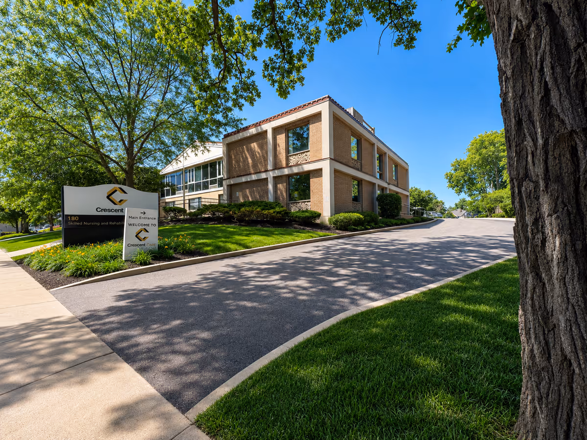 Two-story brick senior living building with curved road, sign, and lush landscaping.