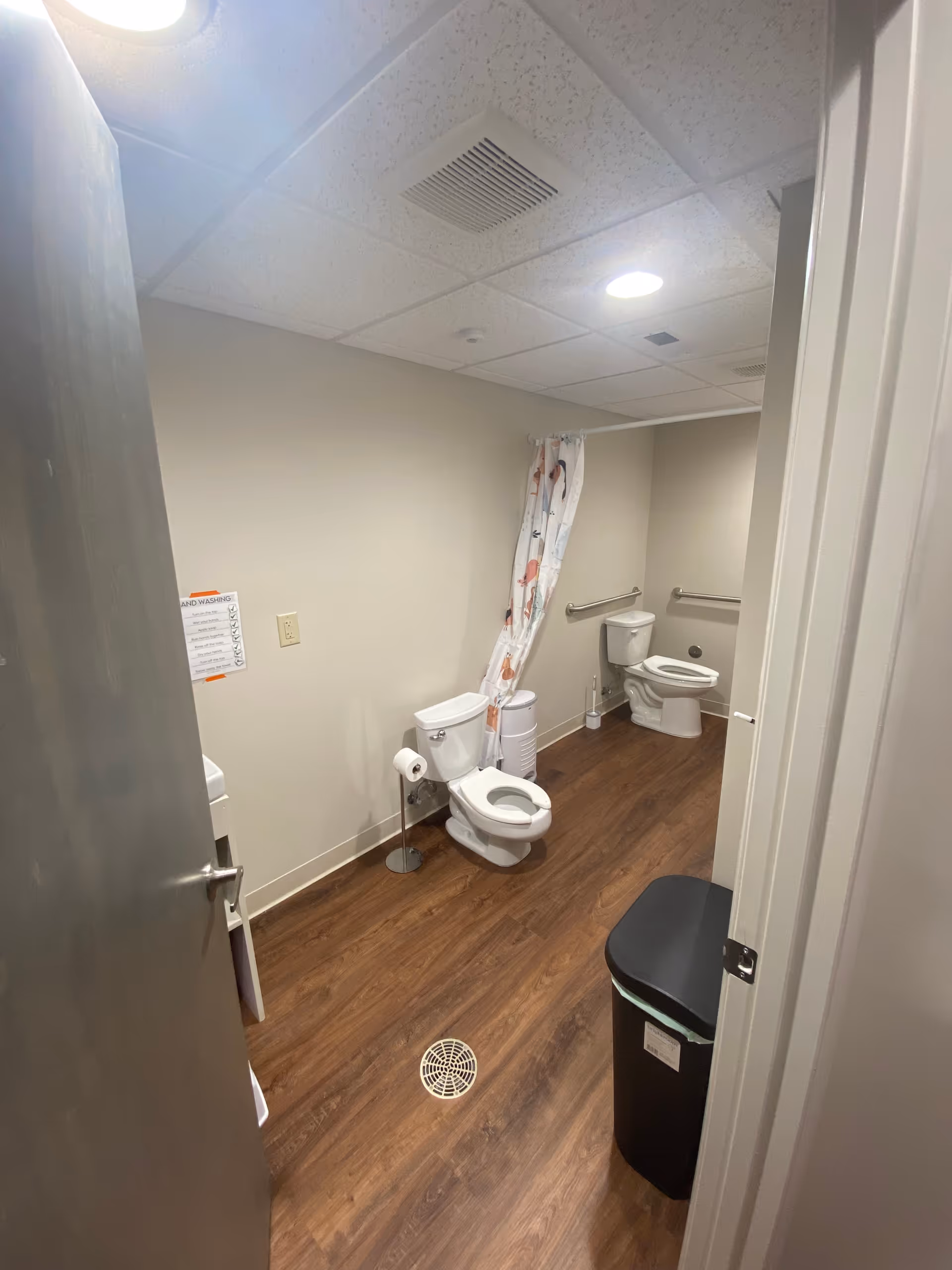Small bathroom with two toilets, shower curtain, grab bars, floor drain, and wood-look flooring.