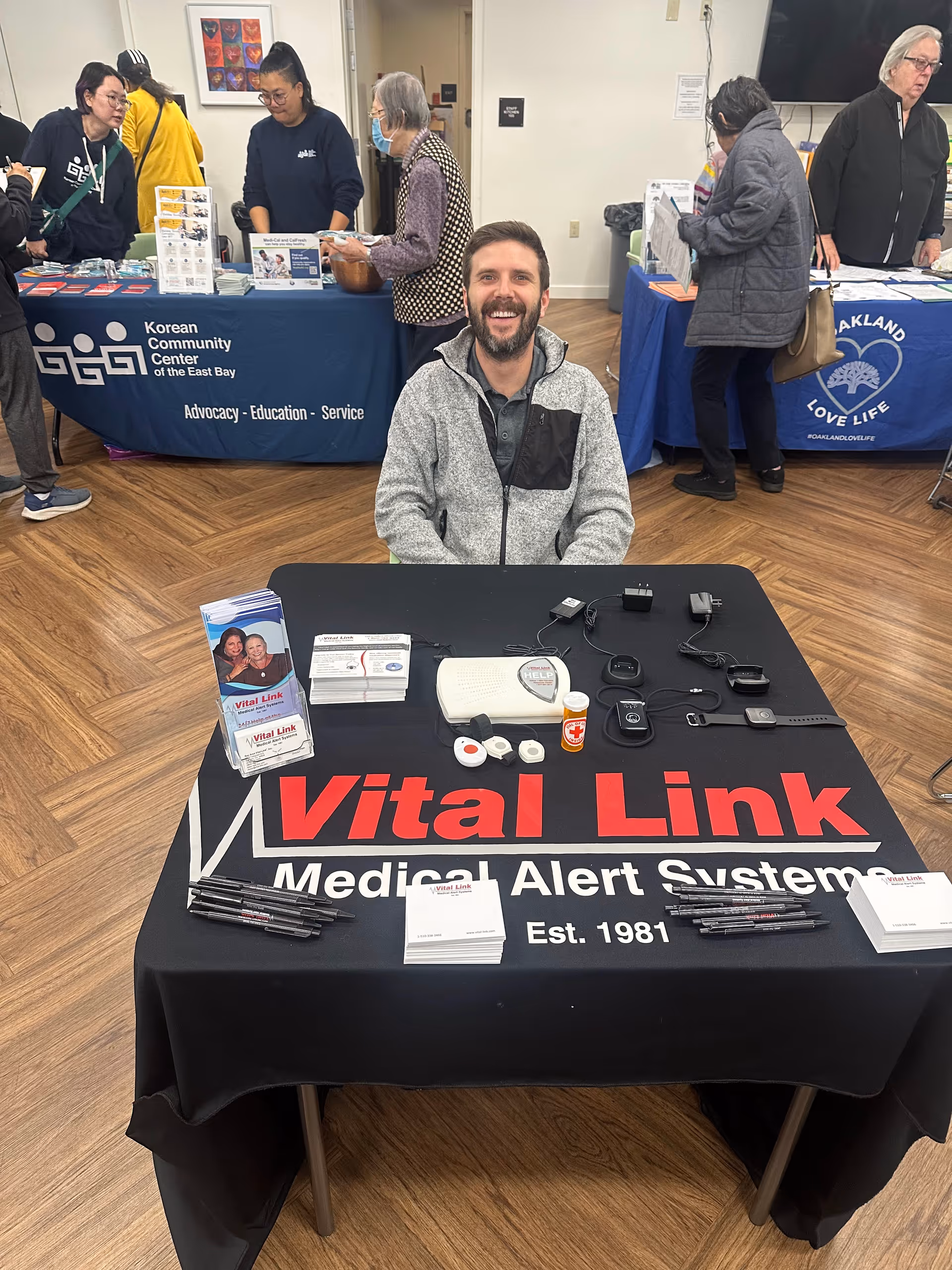 Smiling man sits at a Vital Link medical alert table inside a busy community event.