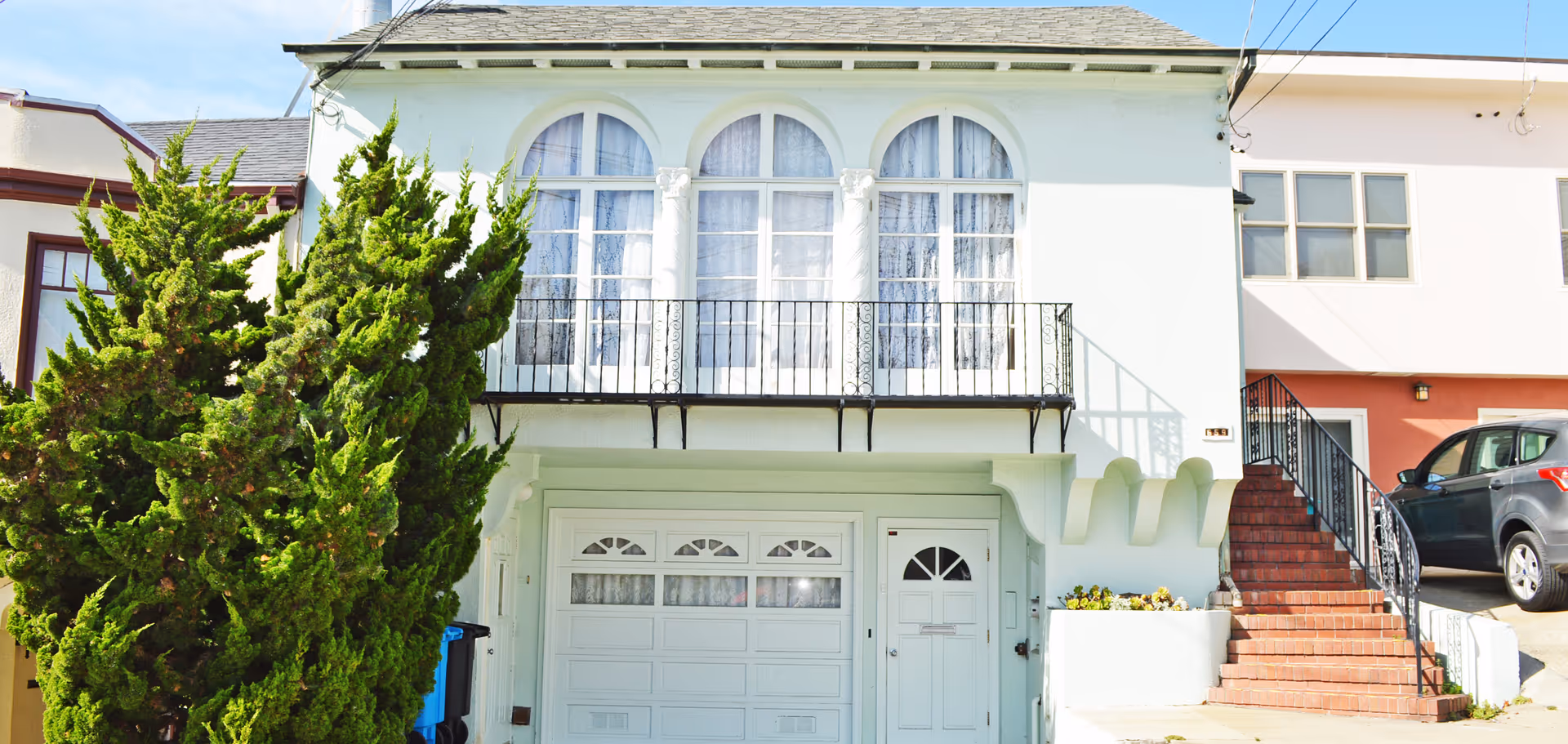 Front exterior of a pale green two-story house with arched windows, a small balcony, garage door, and brick side stairs.