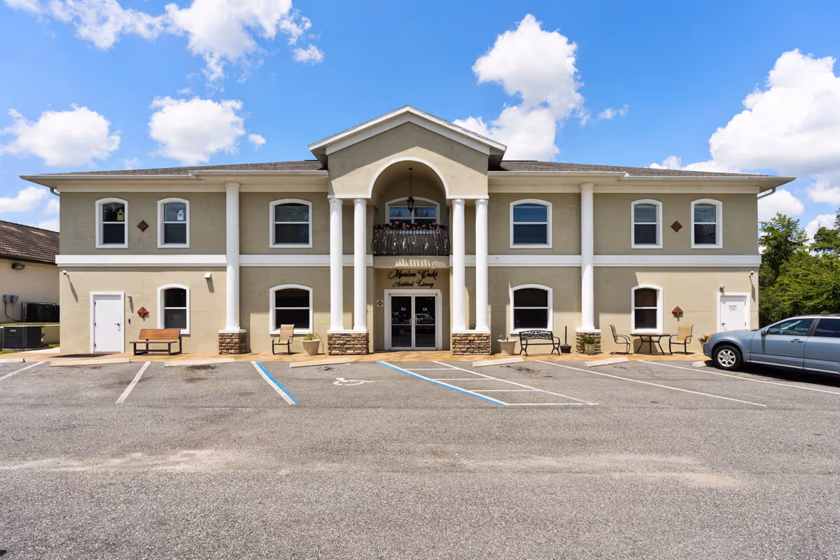Front exterior view of a two-story assisted living facility building with beige walls, white columns, and multiple windows. There is a parking lot in front with marked parking spaces, including a handicapped spot. The entrance has a small balcony above it and outdoor seating with benches and chairs on either side. The sky is blue with scattered clouds.