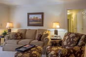 A cozy living room with a beige sofa adorned with patterned cushions, a matching armchair and ottoman with floral upholstery, two table lamps on side tables, a framed landscape painting on the wall, and a hallway leading to other rooms.