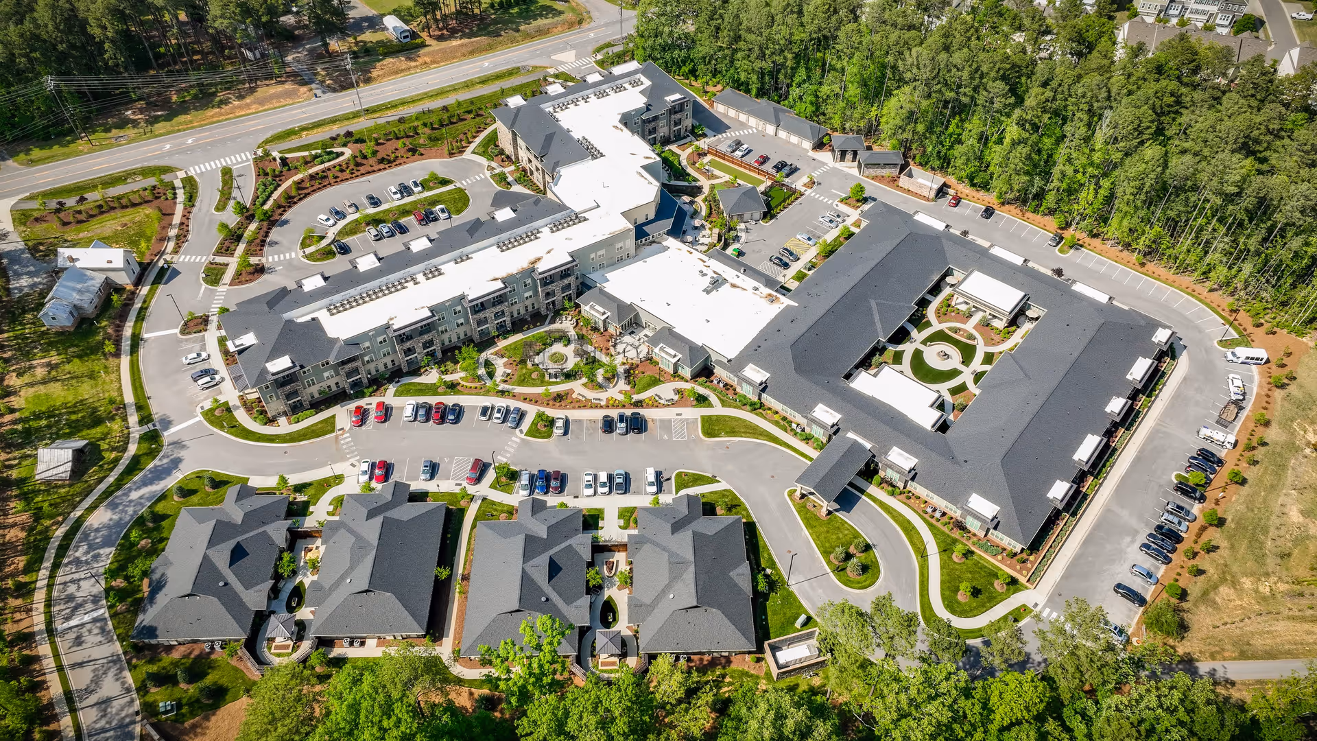 Aerial view of The Reserve at Mills Farm senior living facility showing multiple buildings with dark roofs, parking lots with cars, landscaped gardens, walking paths, and surrounding trees.