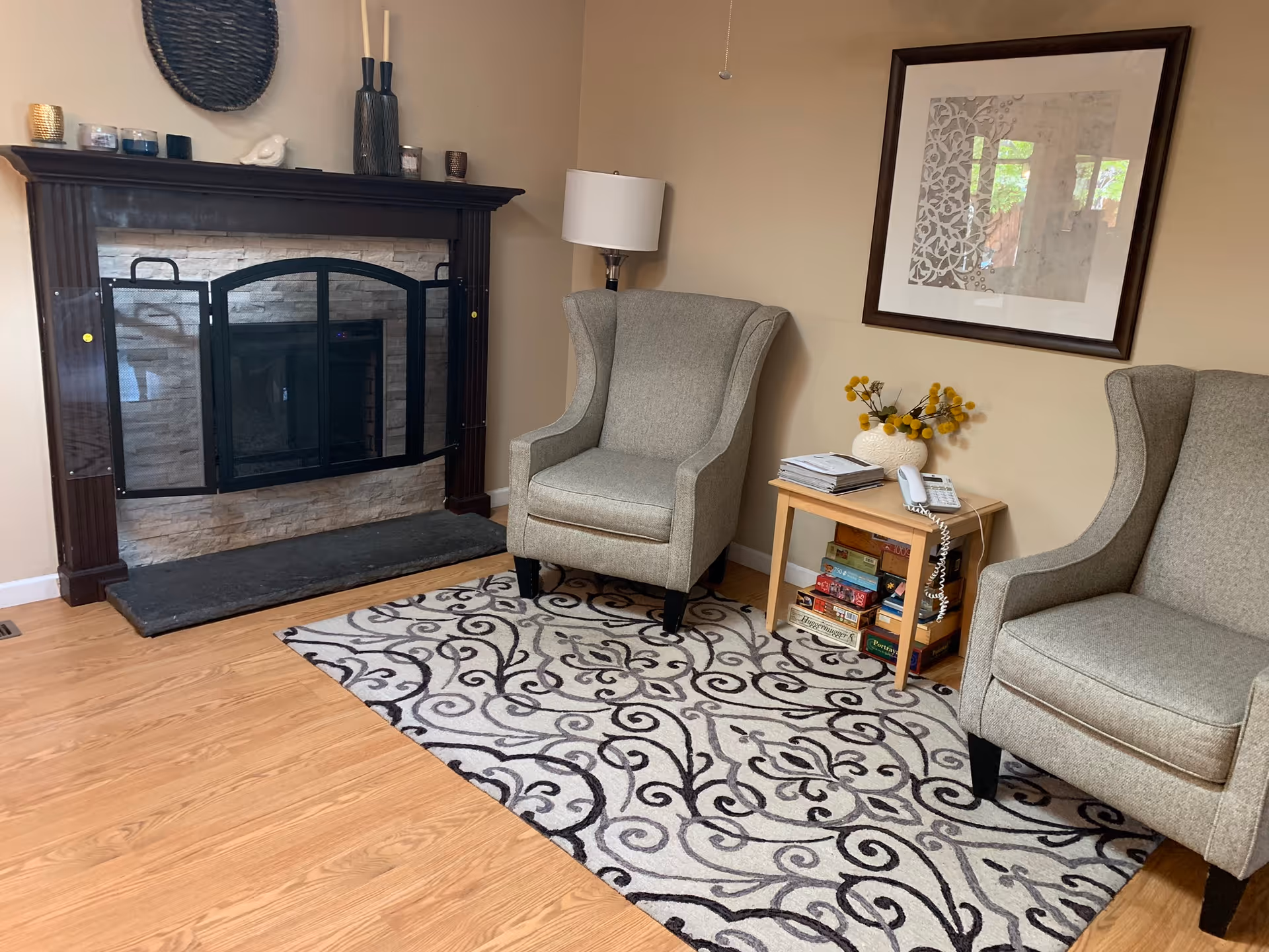 A cozy living room area with two gray upholstered armchairs placed on a patterned rug. Between the chairs is a small wooden side table holding a white vase with yellow flowers, a telephone, and some books and magazines. Behind one chair is a floor lamp, and on the wall above the table is a framed decorative artwork. To the left is a fireplace with a dark wooden mantle decorated with candles and vases.