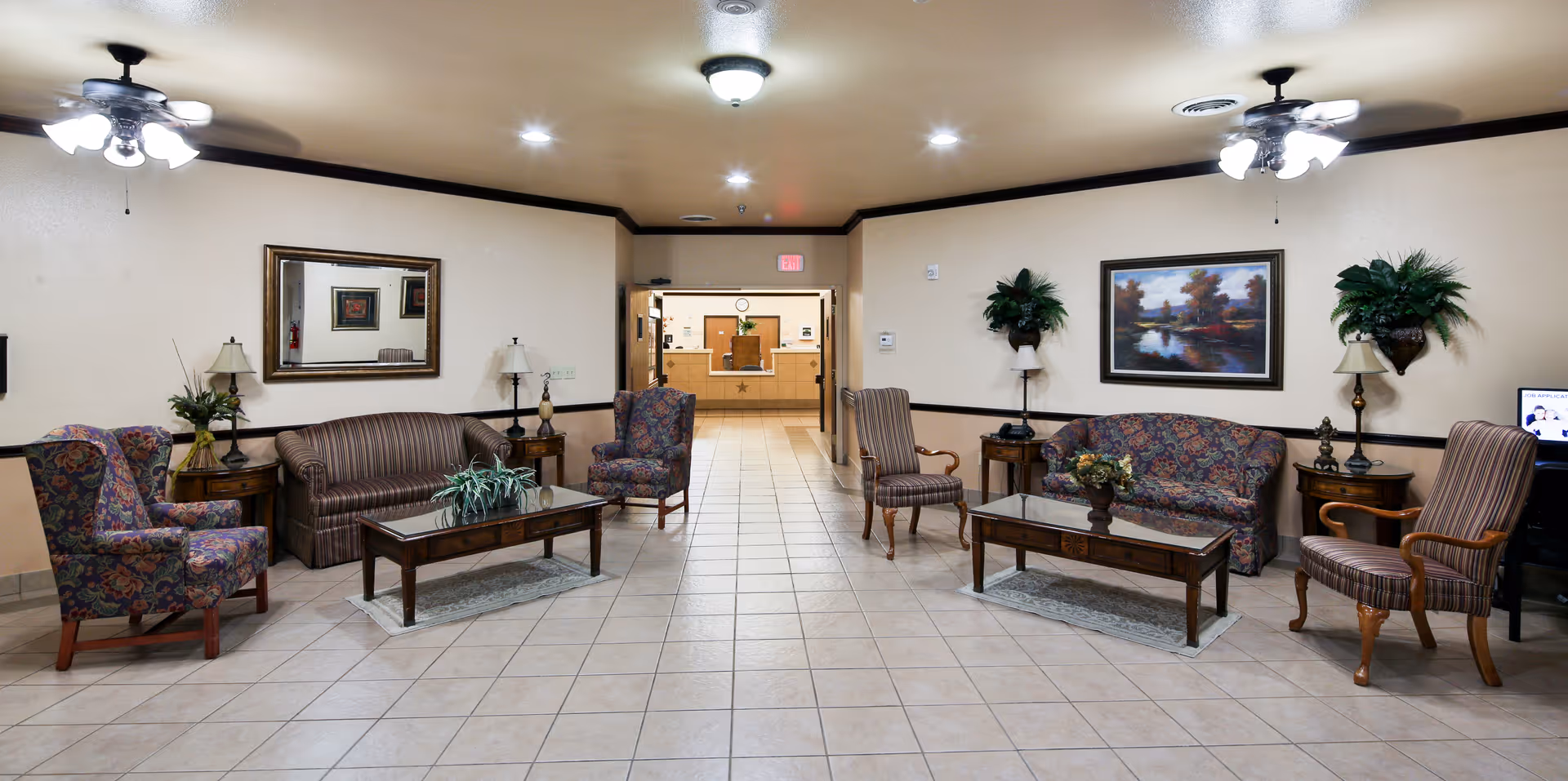 A spacious and well-lit common area with tiled flooring, featuring two seating arrangements each with a sofa, two armchairs, coffee tables, side tables with lamps, and decorative plants. A large mirror and a framed painting hang on the walls. Ceiling fans with lights are mounted on the ceiling. In the background, a reception desk area is visible through an open doorway.