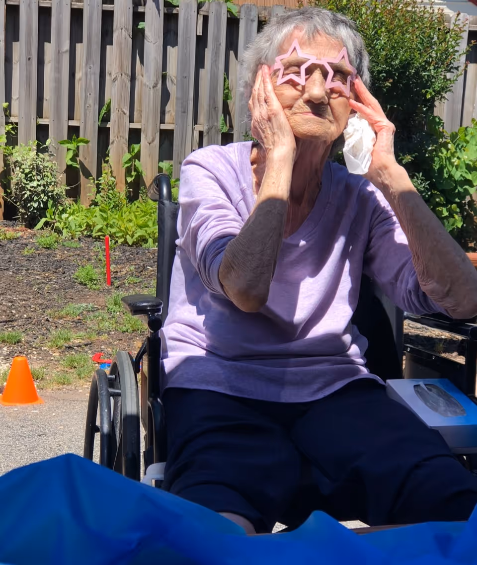 An elderly woman sitting in a wheelchair outdoors, wearing a light purple long-sleeve shirt and dark pants, playfully holding pink star-shaped sunglasses to her face. She is in a garden area with a wooden fence and greenery in the background.