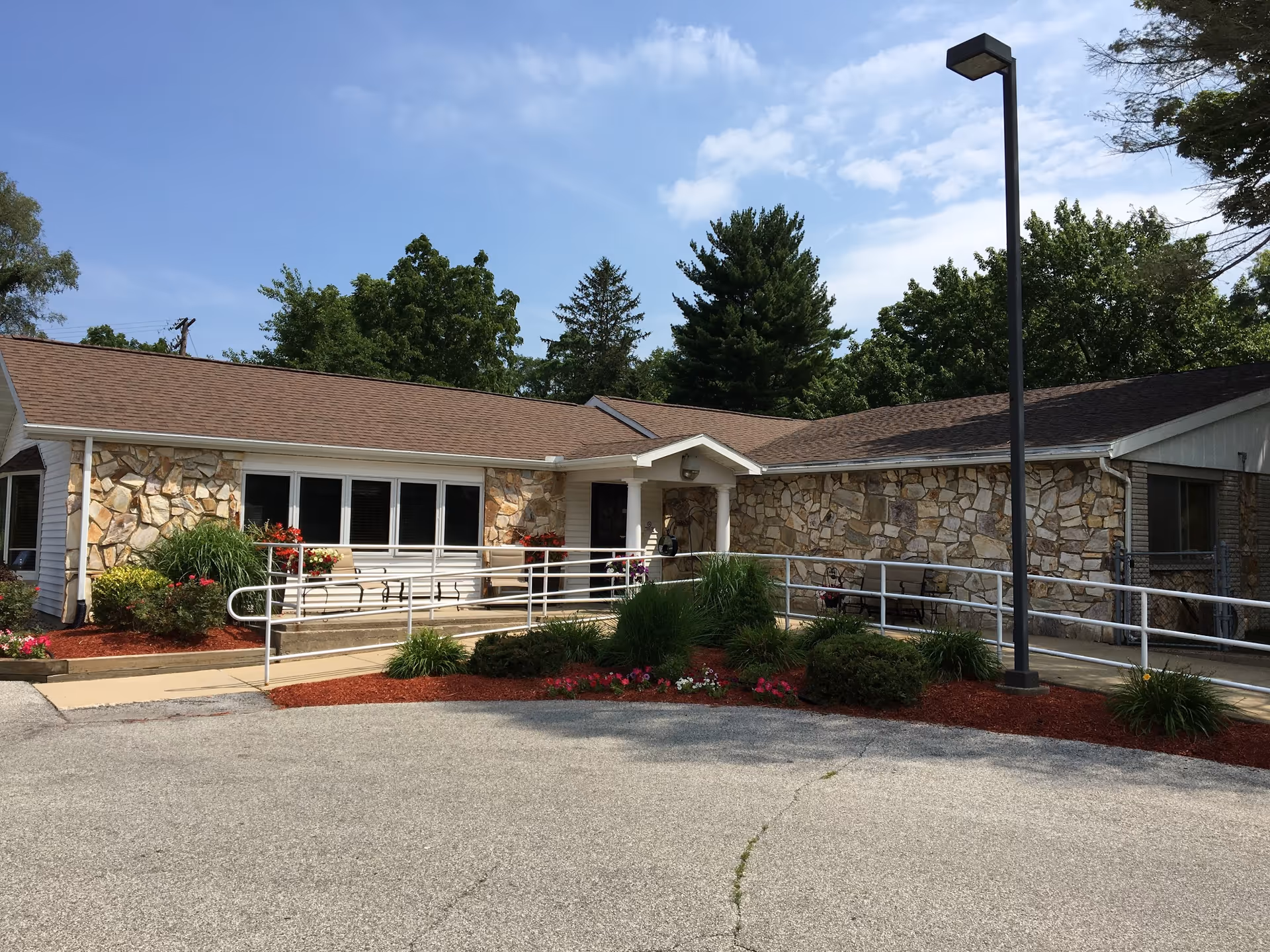 Single-story stone-faced senior care building front with a wheelchair ramp, landscaped flower beds, and a tall lamp post under a blue sky.