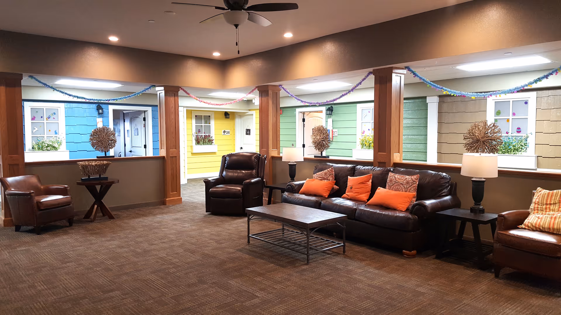Interior common area with brown leather chairs and sofa with orange pillows, a wooden coffee table, and side tables with lamps. The walls have large open windows showing colorful cottage-style doors and windows decorated with flowers and festive garlands hanging from the ceiling.