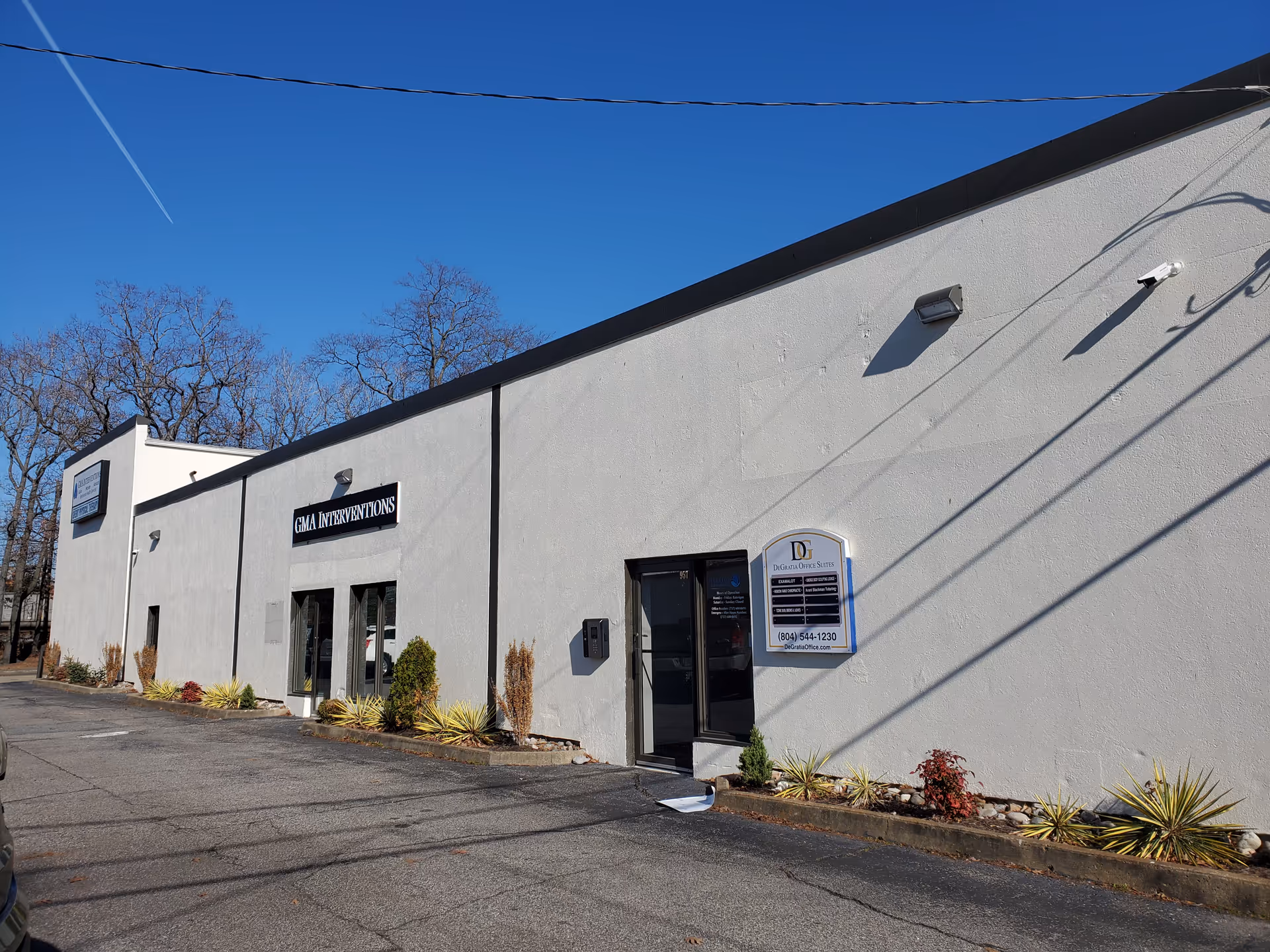 Grey commercial building exterior with doors, windows, a parking lot and landscaping.