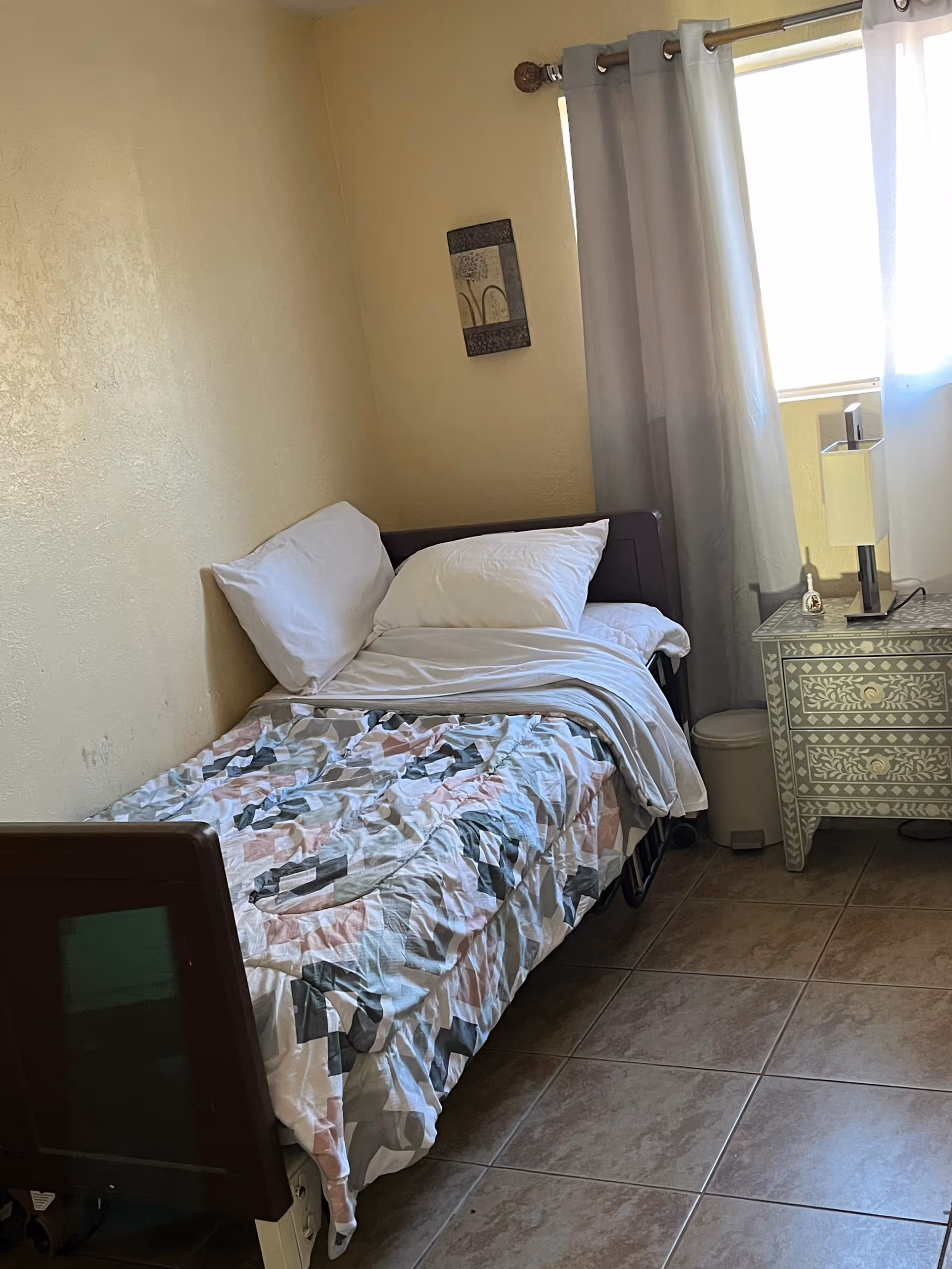 Small bedroom with a single bed, quilt, pillows, nightstand, lamp, and a window with curtains.