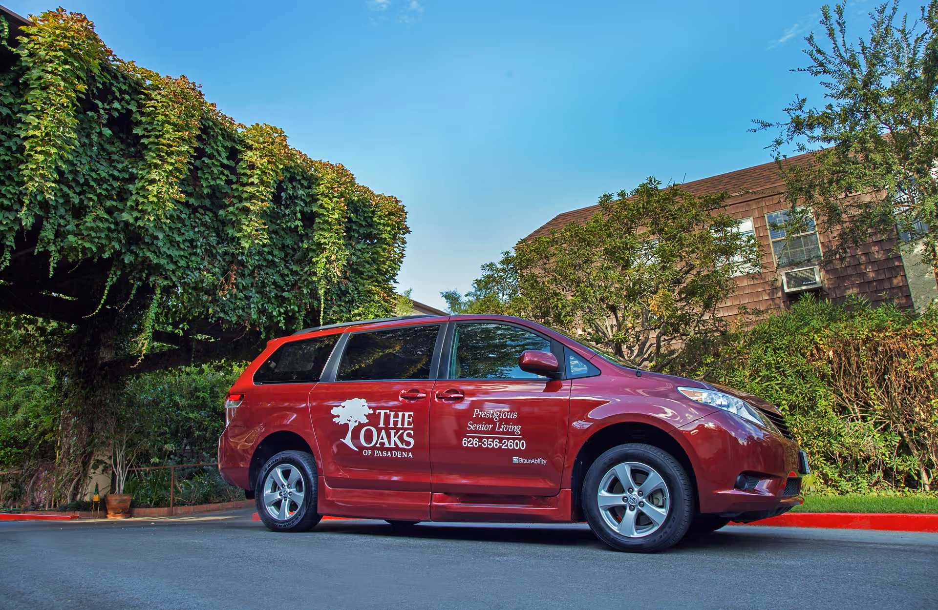A red minivan parked on a driveway outside a building with trees and bushes around. The minivan has the logo and text for The Oaks of Pasadena, a senior living facility, including a phone number and the phrase 'Prestigious Senior Living'.