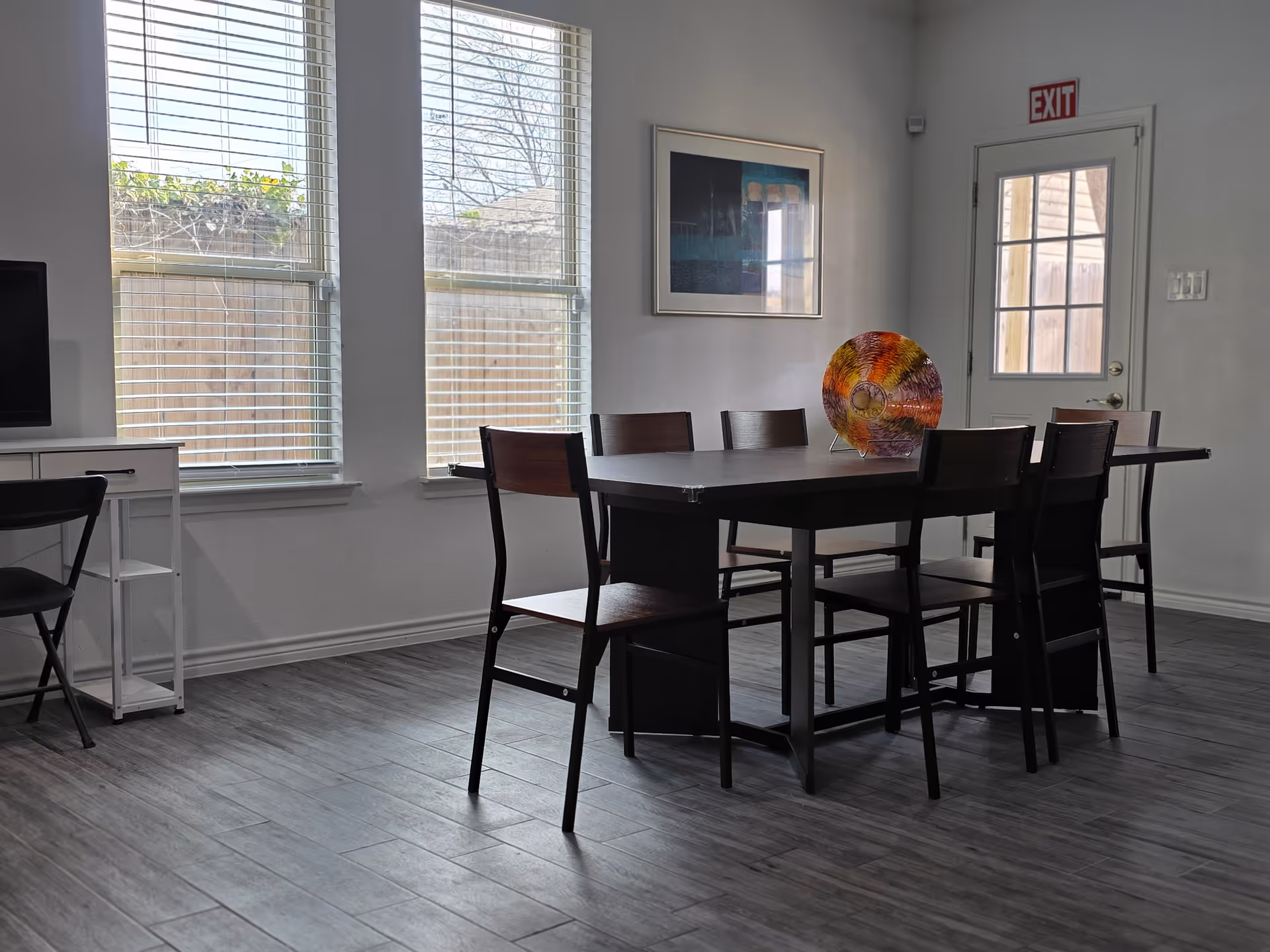 A dining room with a long dark table, chairs, windows, and an exit door.
