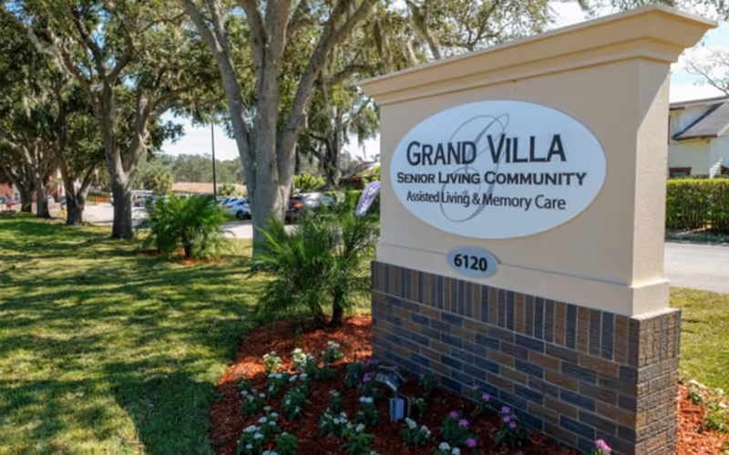 Entrance sign reading 'Grand Villa Senior Living Community' with landscaped flower beds and trees along a driveway.