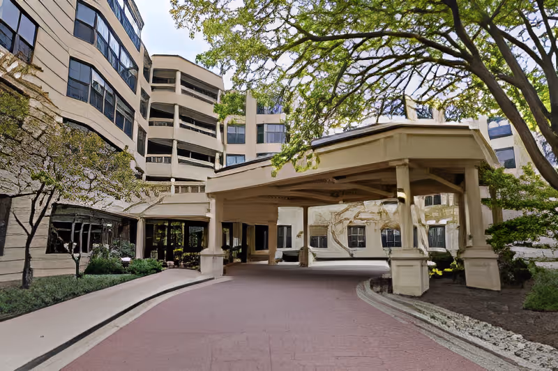 Covered porte-cochère and circular driveway at the entrance of a multi-story senior living building surrounded by trees.