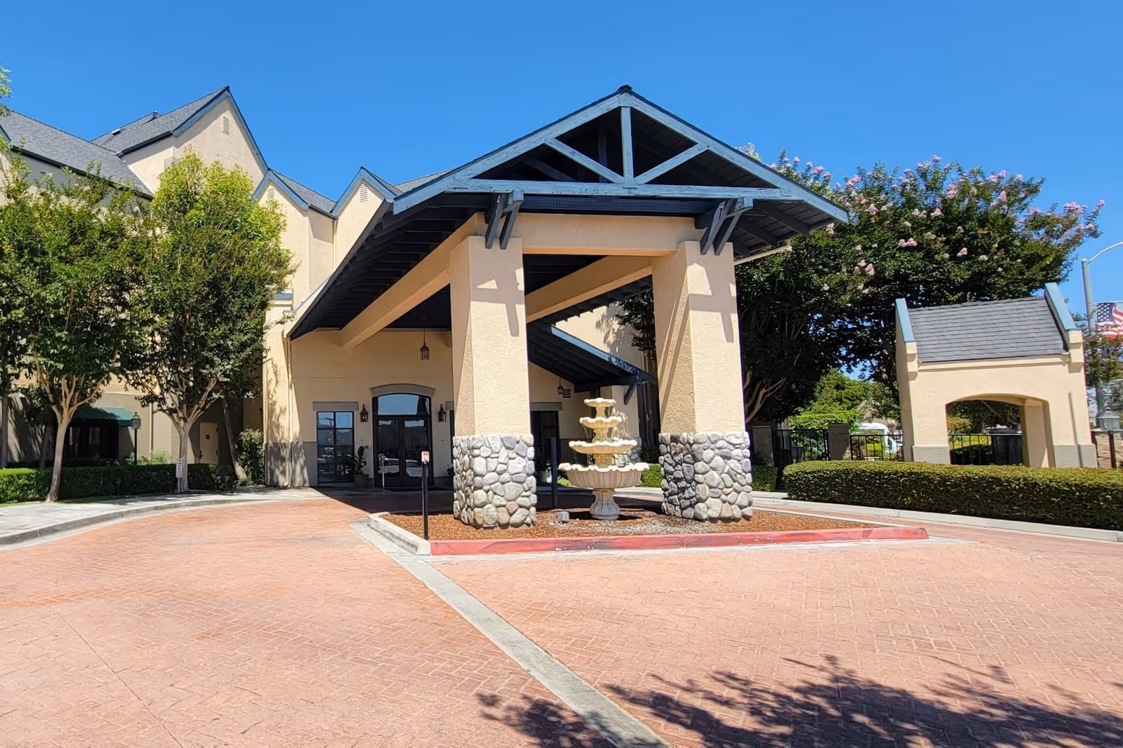 Front entrance of Westminster Terrace showing a covered porte-cochere with stone columns, a tiered fountain, and landscaped driveway under a clear blue sky.