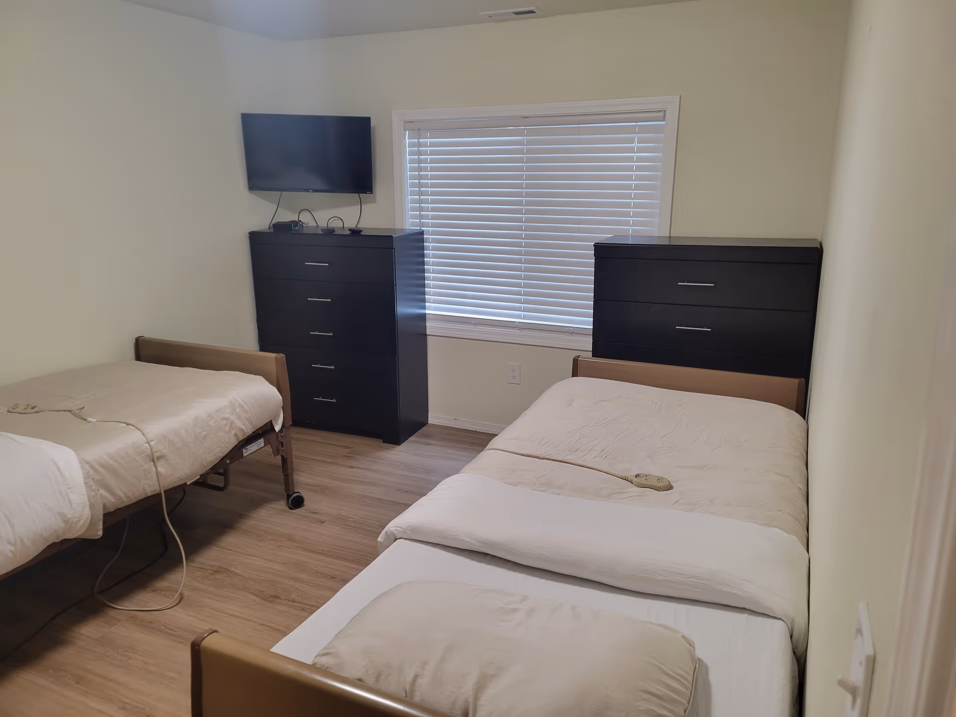 Two single beds with beige bedding, dark dressers, and a window in a small shared bedroom.