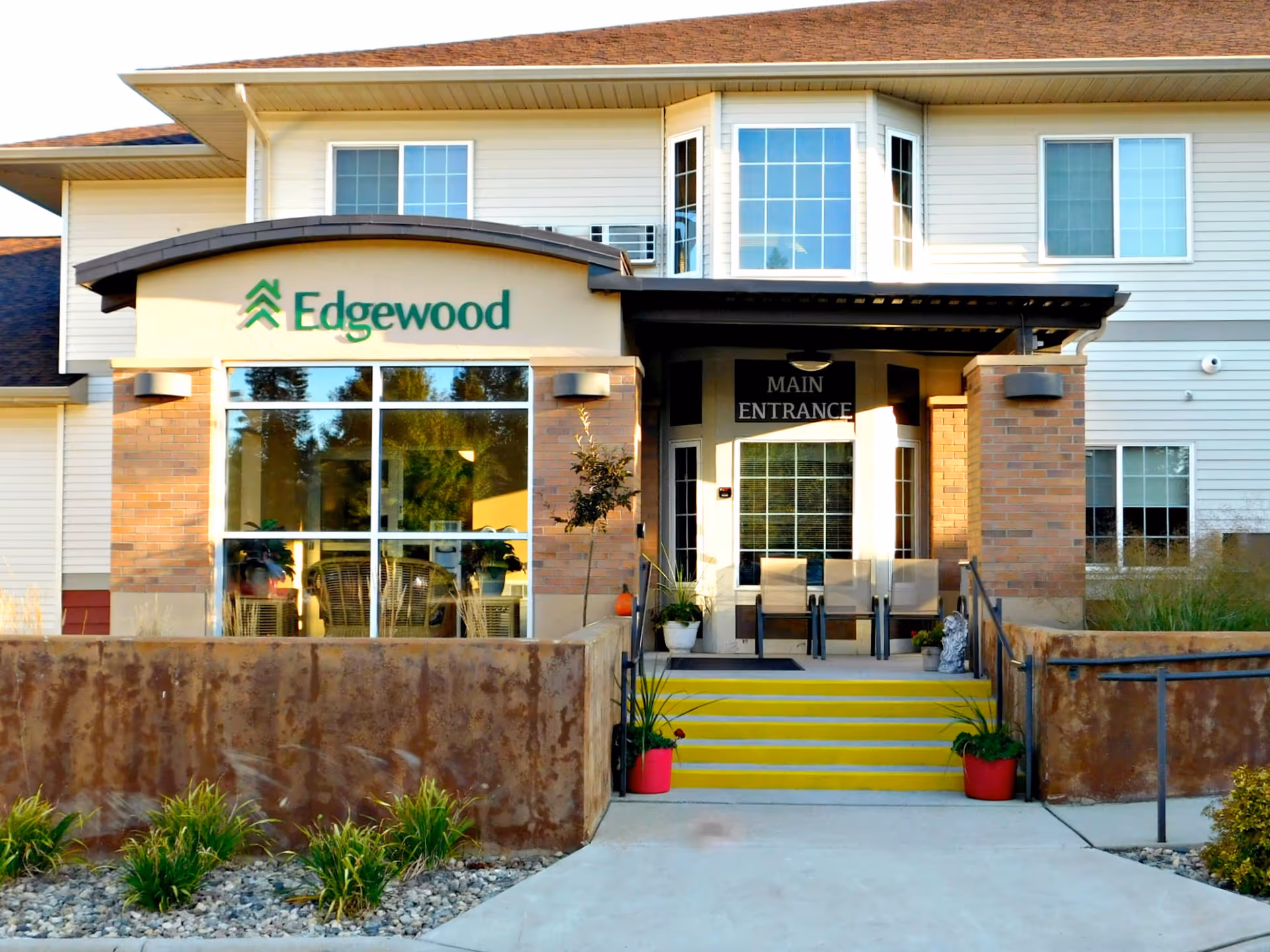 Front exterior view of the Edgewood Watertown facility showing the main entrance with yellow steps, potted plants, and a covered porch area. The building has large windows and a sign with the Edgewood logo above the entrance.
