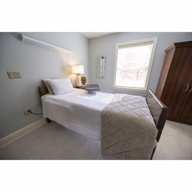 A small hospital-style bedroom with a single bed, nightstand, lamp, and wardrobe.