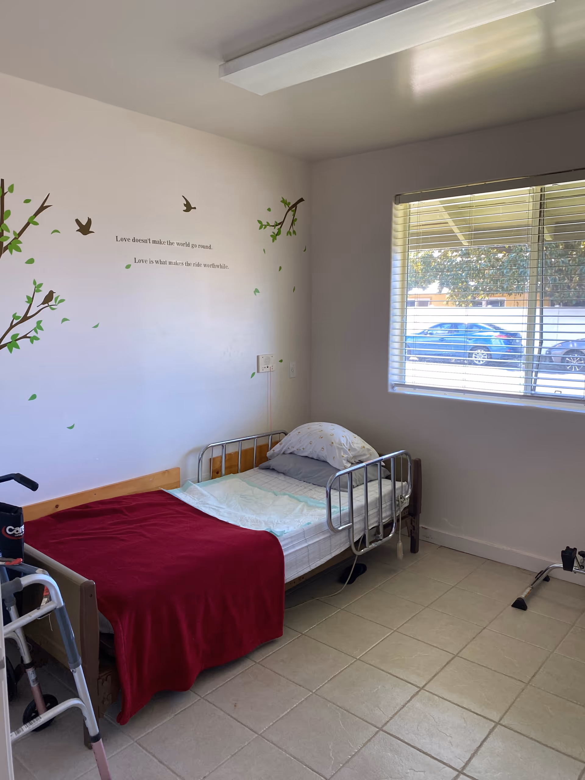 A small resident bedroom with a single bed, wall decals, a window, and a walker nearby.