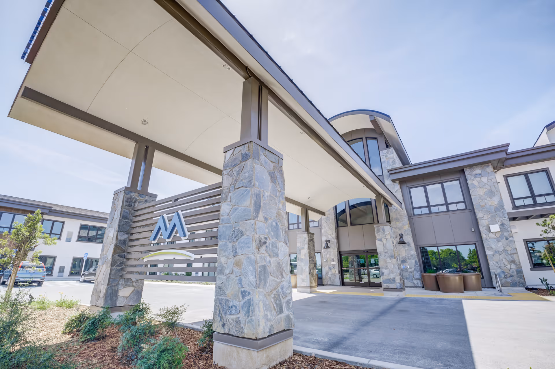 Entrance of a senior living facility with stone pillars and a covered driveway. The building has large windows and a modern design with a clear blue sky in the background.