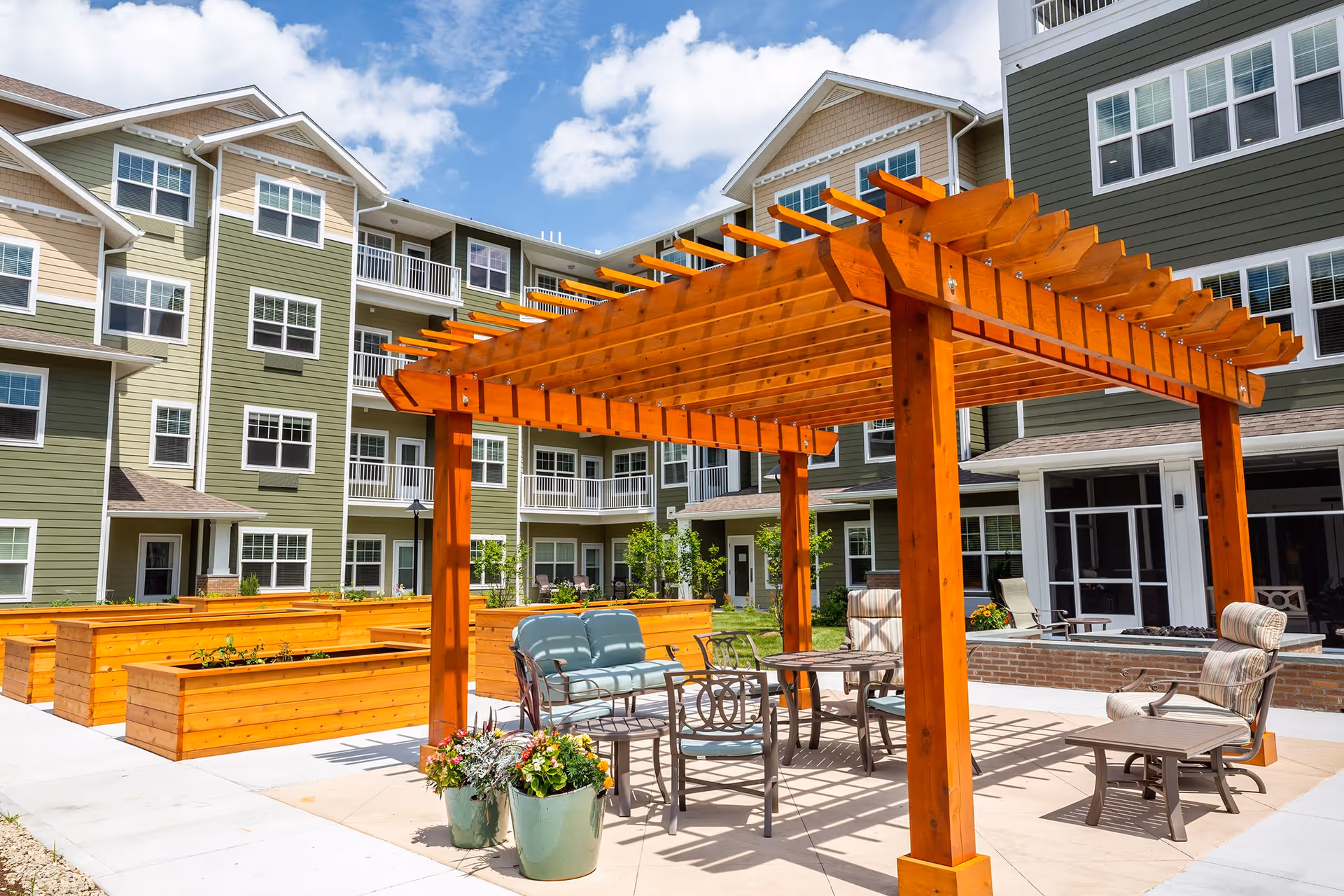 Outdoor patio area at Camellia Gardens Gracious Retirement Living featuring a wooden pergola with seating including cushioned chairs and a loveseat, surrounded by raised garden beds and a multi-story residential building in the background under a partly cloudy sky.