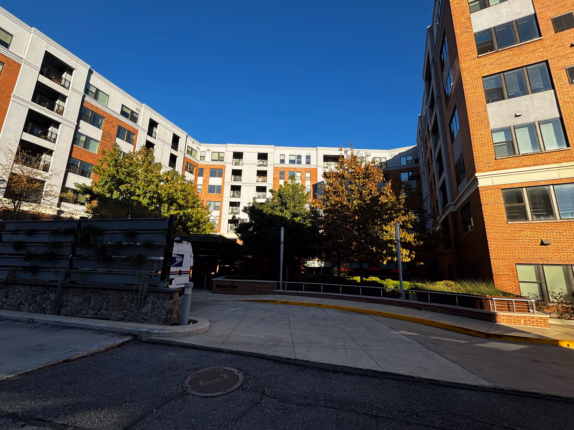 Exterior view of a multi-story senior living facility building with a mix of brick and light-colored walls, several windows, and balconies. There are trees and shrubs in front of the building, a curved driveway, and a USPS delivery van parked near the entrance under a covered area. The sky is clear and blue.