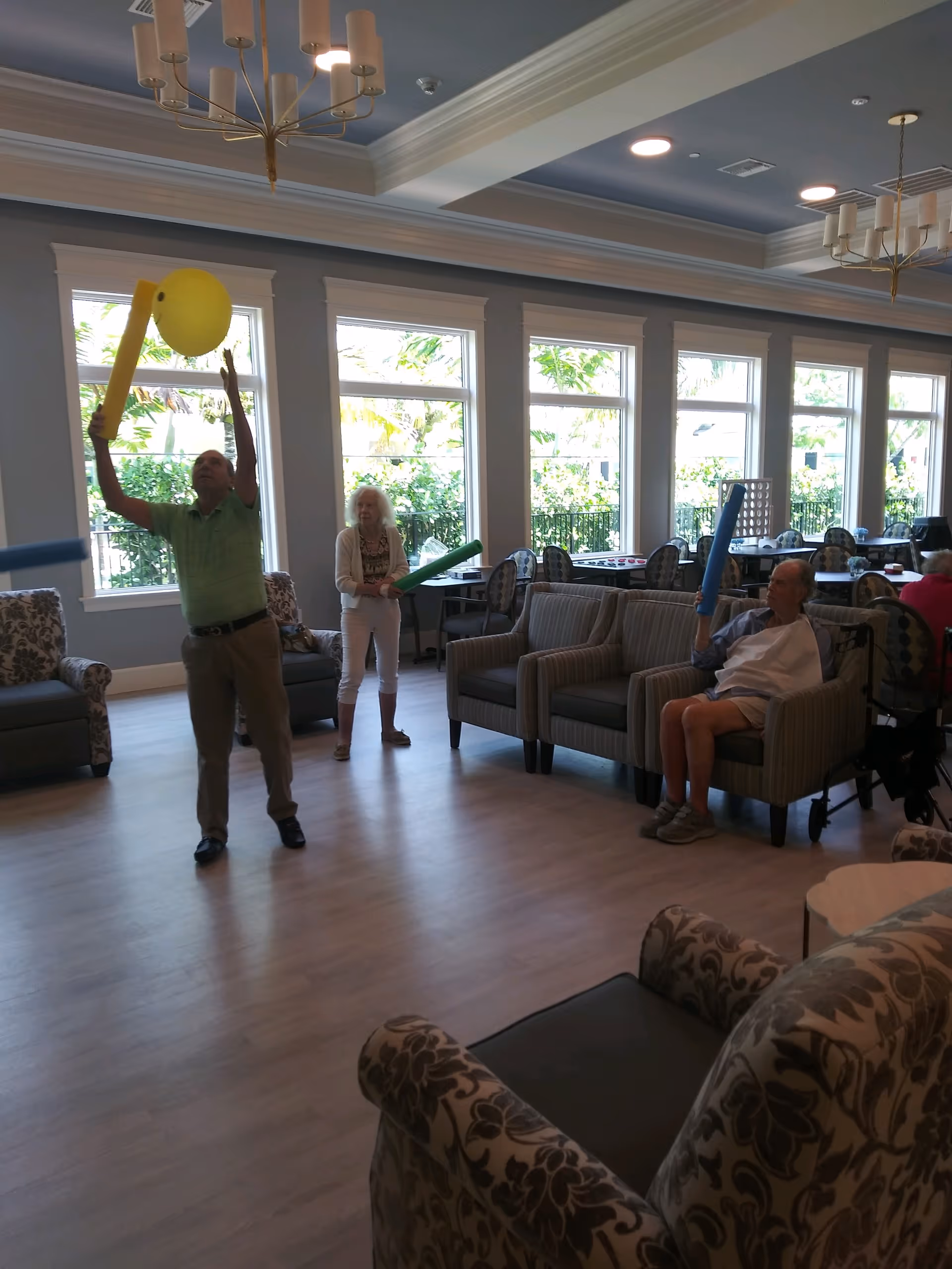 A group of elderly people in a spacious, well-lit common room with large windows. One man is standing and hitting a large yellow balloon with a foam noodle, while a woman stands nearby holding a green foam noodle. Another man is seated on a couch holding a blue foam noodle. The room has comfortable armchairs, sofas, and chandeliers hanging from the ceiling.
