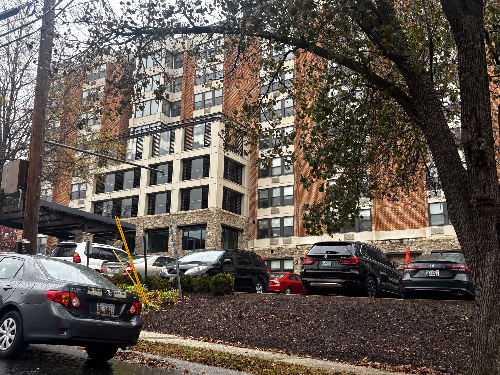 Exterior front of a multi-story brick residential building with parked cars and a tree in the foreground.