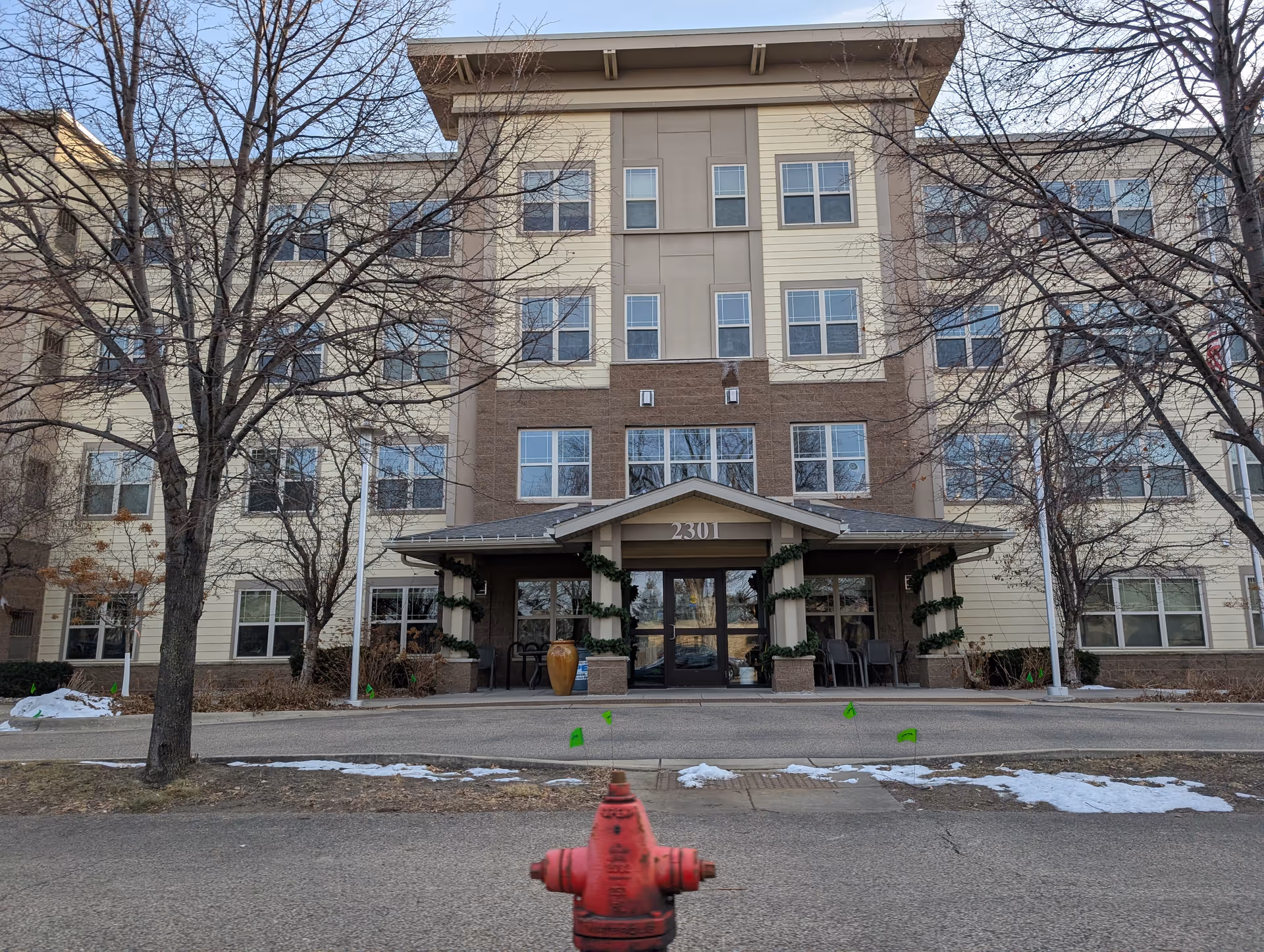 Front exterior view of a multi-story senior living facility building with the address number 2301 above the entrance. The entrance is decorated with garlands wrapped around the pillars. Leafless trees and patches of snow are visible in the foreground along with a red fire hydrant near the street.