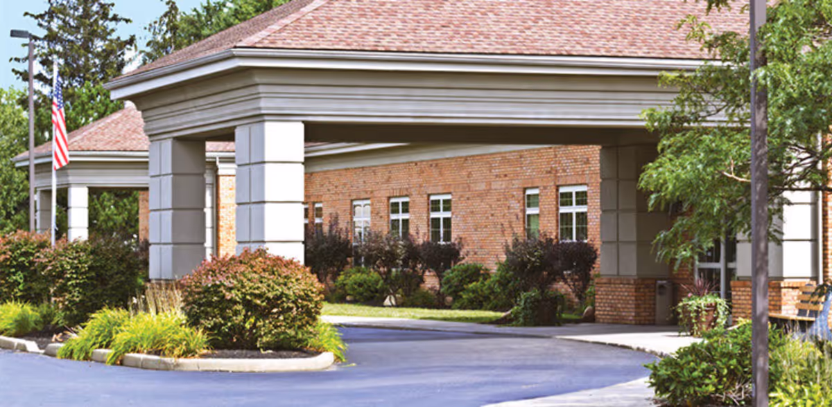 Entrance of a brick building with a covered driveway supported by large white columns, surrounded by green bushes and trees, with an American flag on a flagpole to the left.