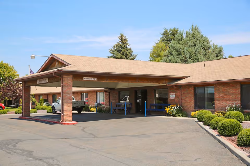 Brick senior care facility with a covered entrance, vehicles, and landscaped shrubs.