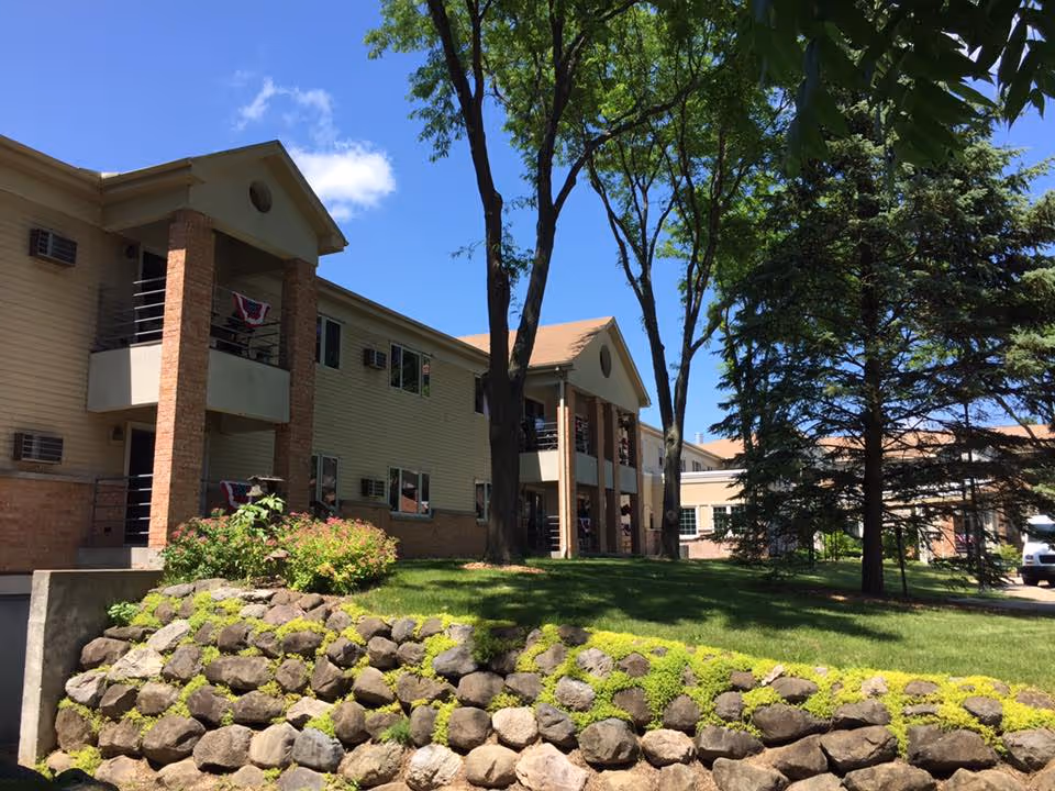 Exterior view of Trinity Pines Retirement Center with a stone retaining wall, trees, and green lawn.
