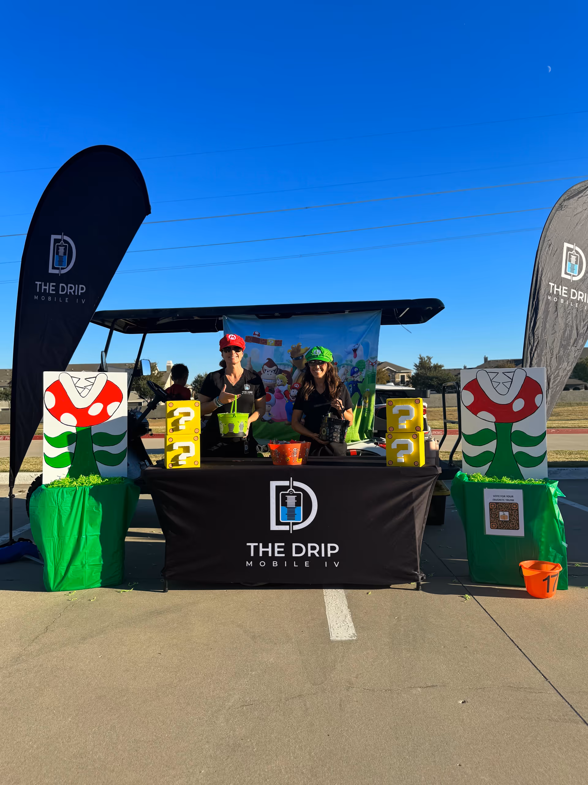 Two staff members operate The Drip Mobile IV booth outdoors at a sunny event.
