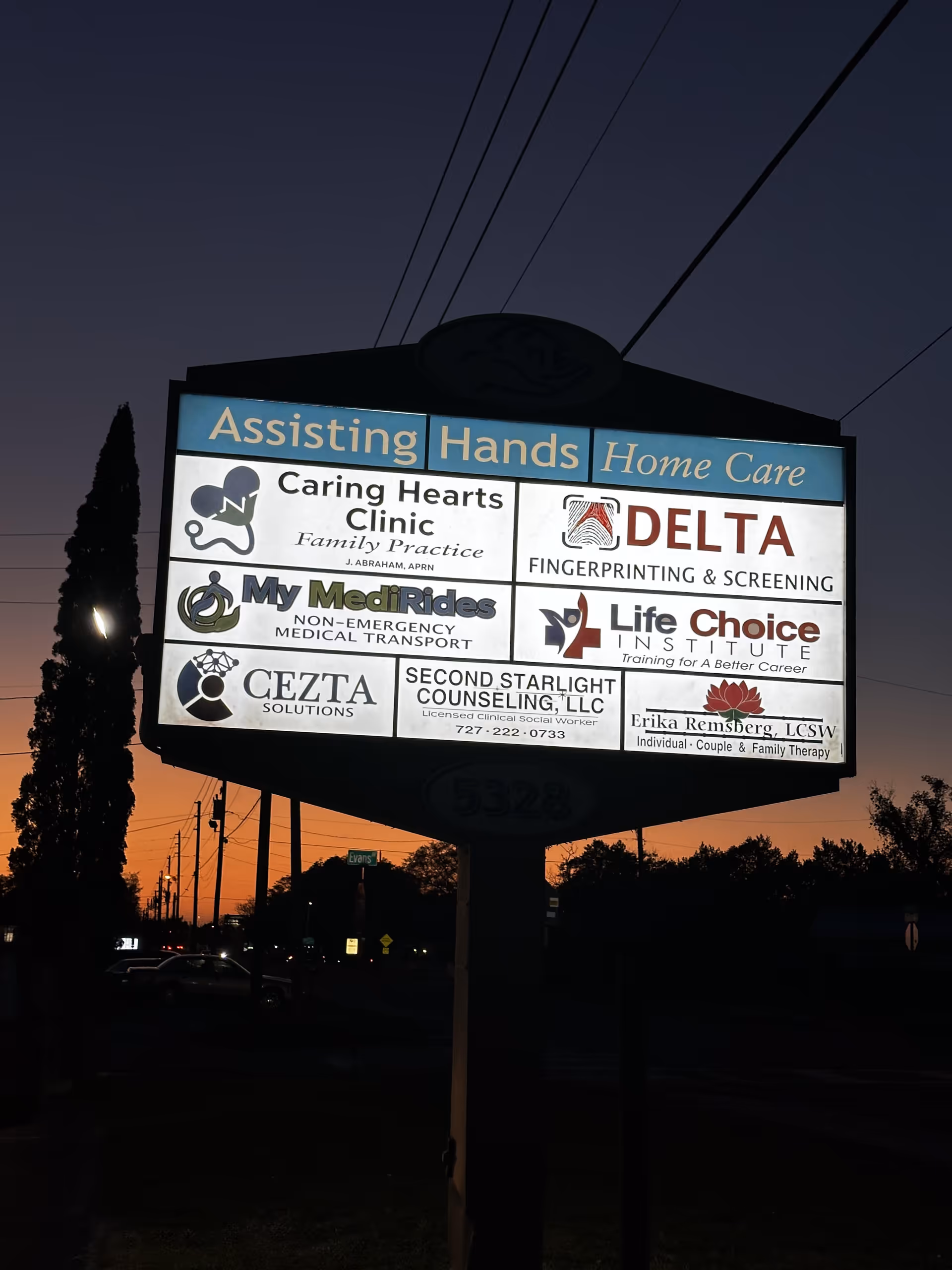 Illuminated exterior sign for Assisting Hands Home Care displaying multiple logos at dusk
