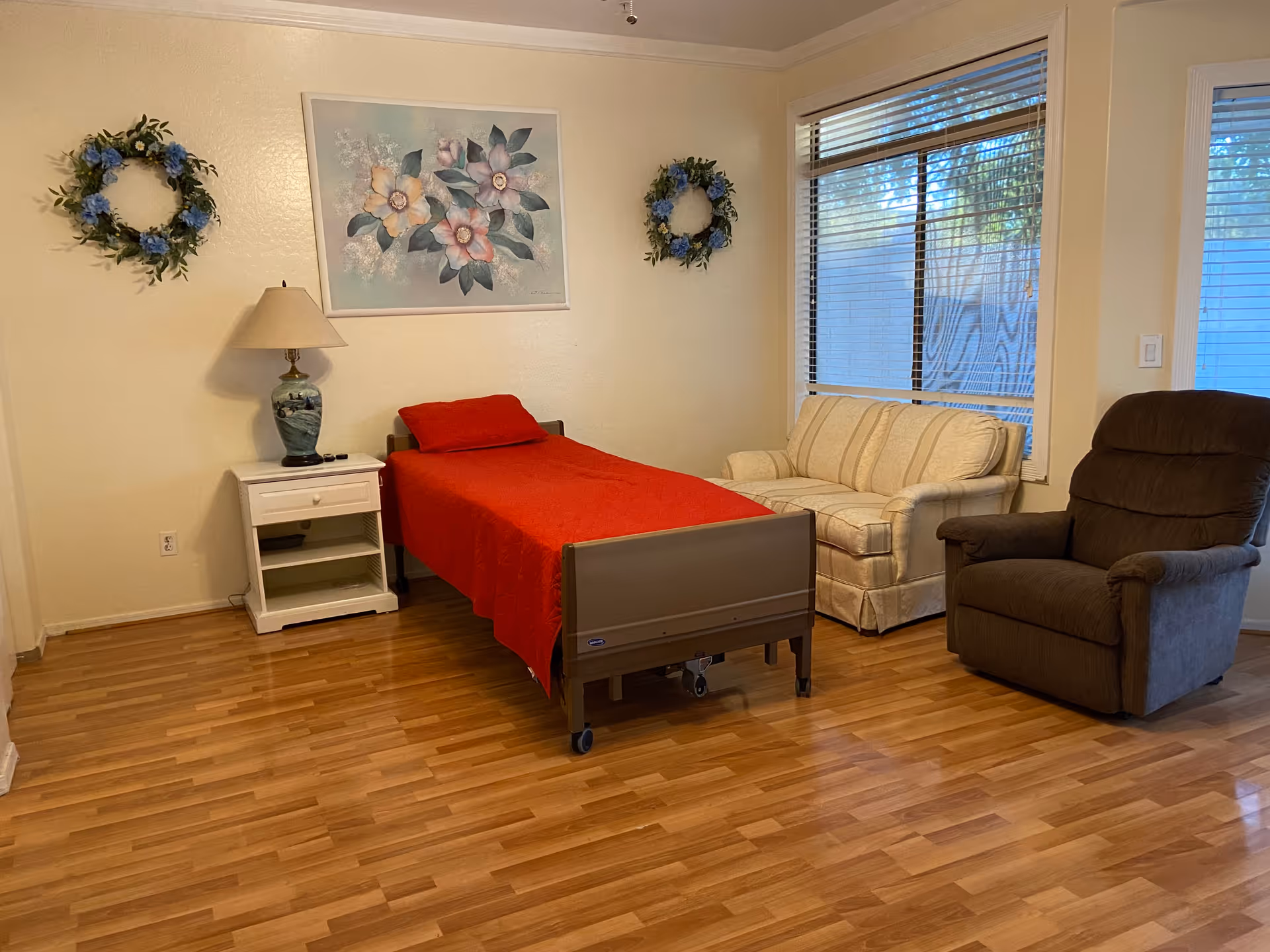 A furnished senior facility bedroom with a red bedspread, nightstand, sofa, and recliner.