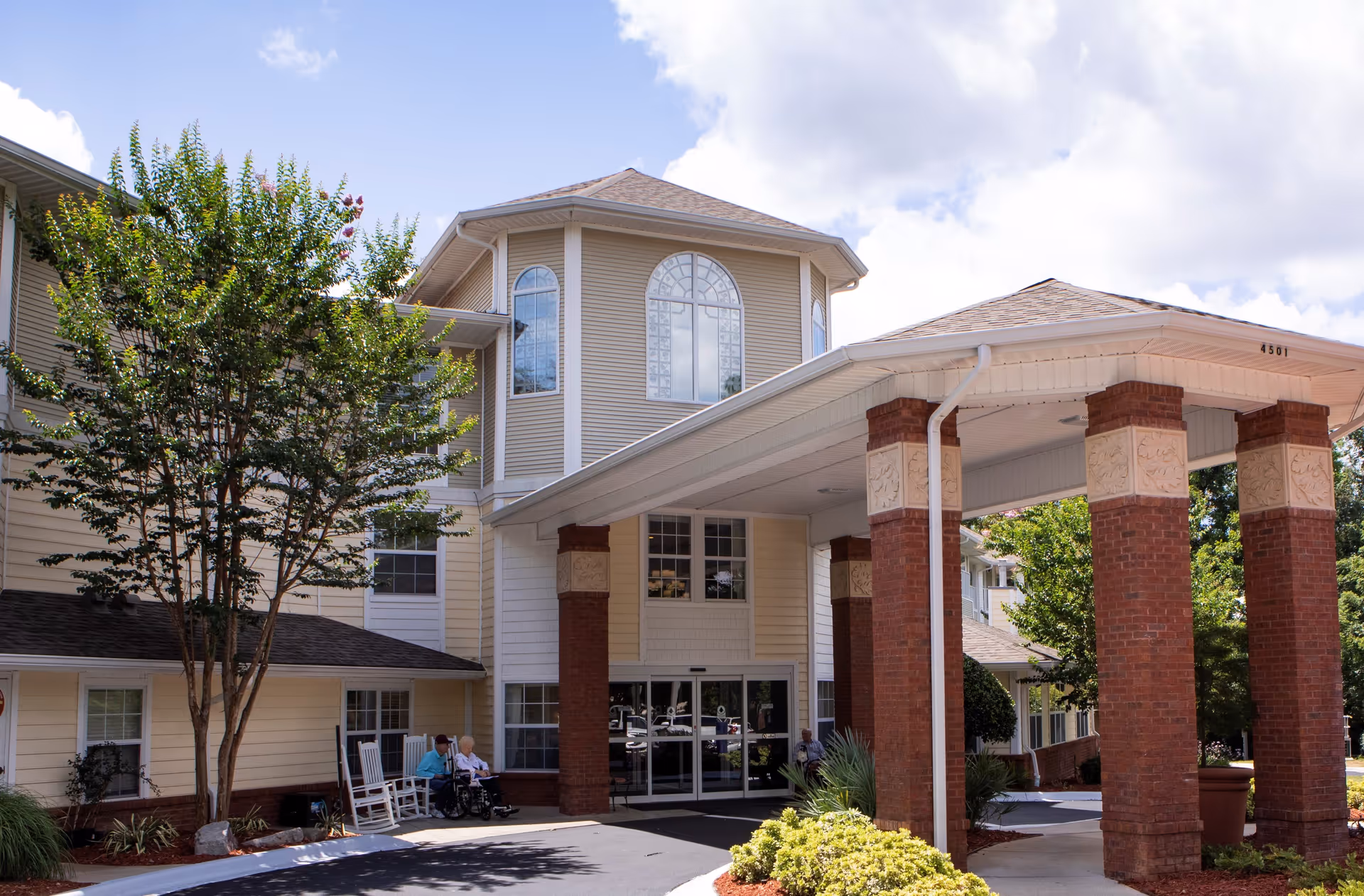 Exterior view of Allegro Senior Living facility in Tallahassee, FL, showing the entrance with a covered driveway supported by brick columns, a tree, shrubs, and two elderly people sitting on rocking chairs near the entrance.