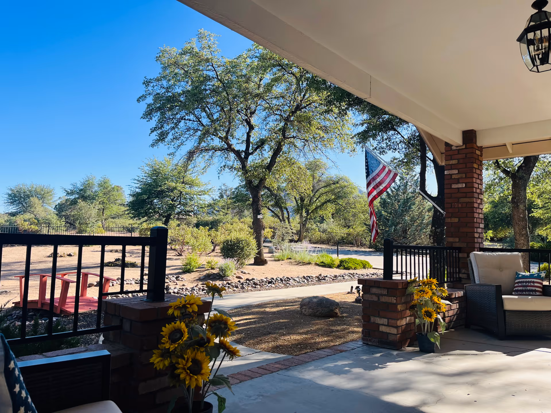 Outdoor covered porch with brick columns, seating, sunflowers, and an American flag overlooking a sunny landscape.