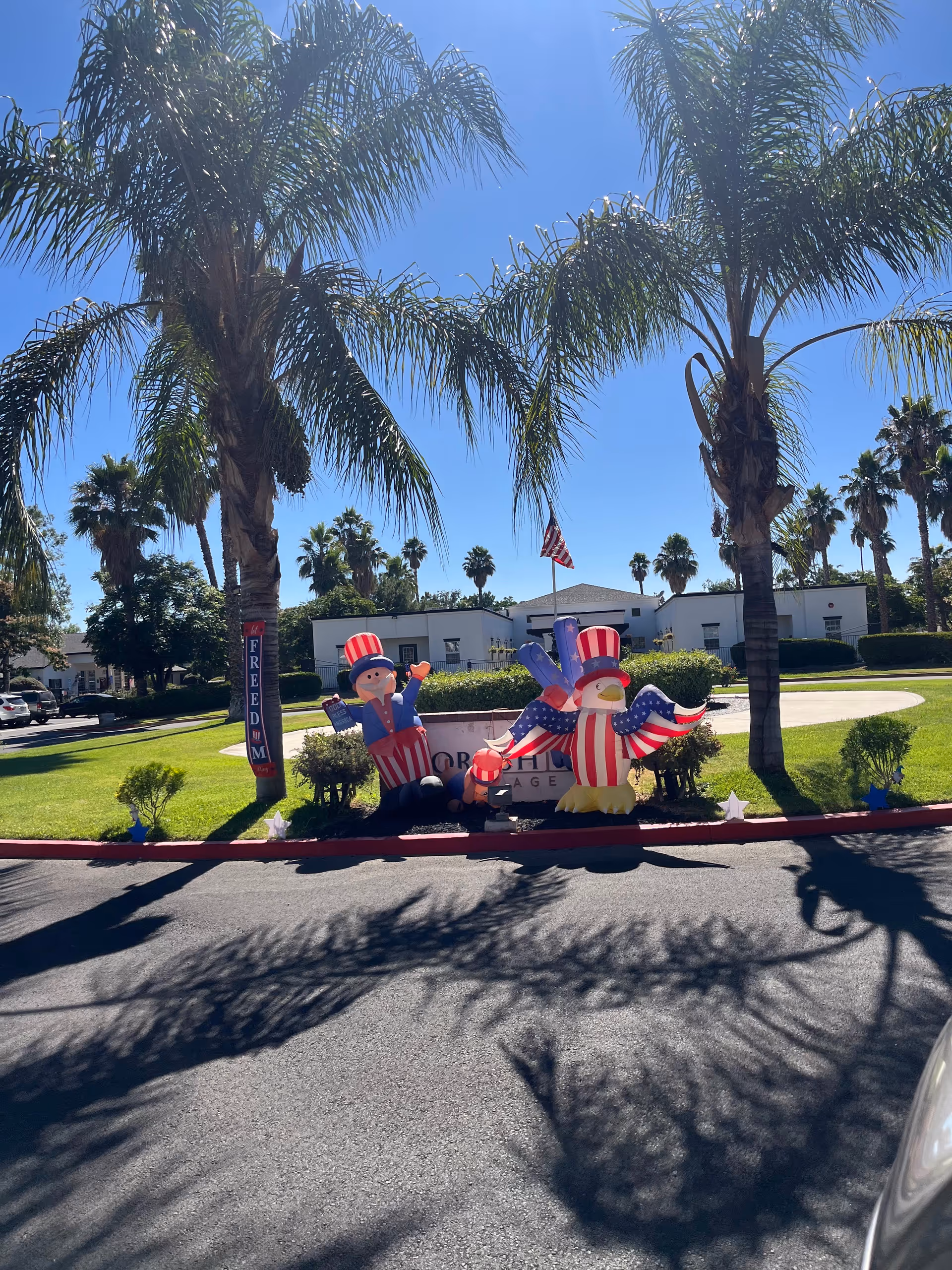 Sunny exterior of Yorkshire Village senior living with palm trees and patriotic inflatables in front yard.