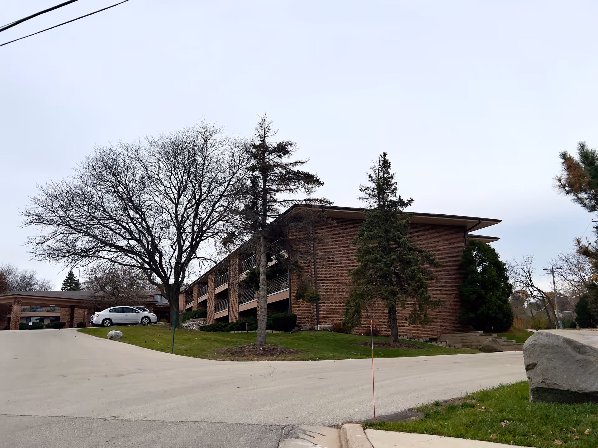 Brick two-story senior living building with balconies, trees, a driveway and a parked white car.