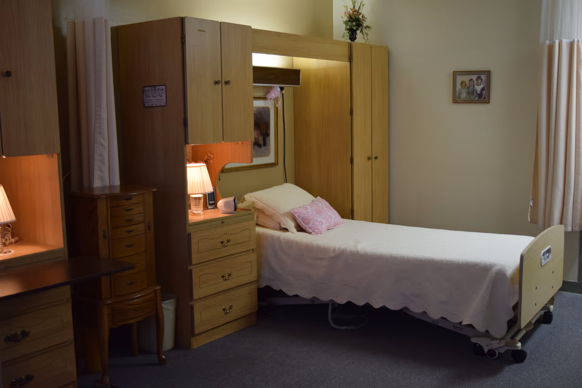 Senior living facility bedroom with wooden wardrobes, a bed, dresser, nightstand, and lamp.