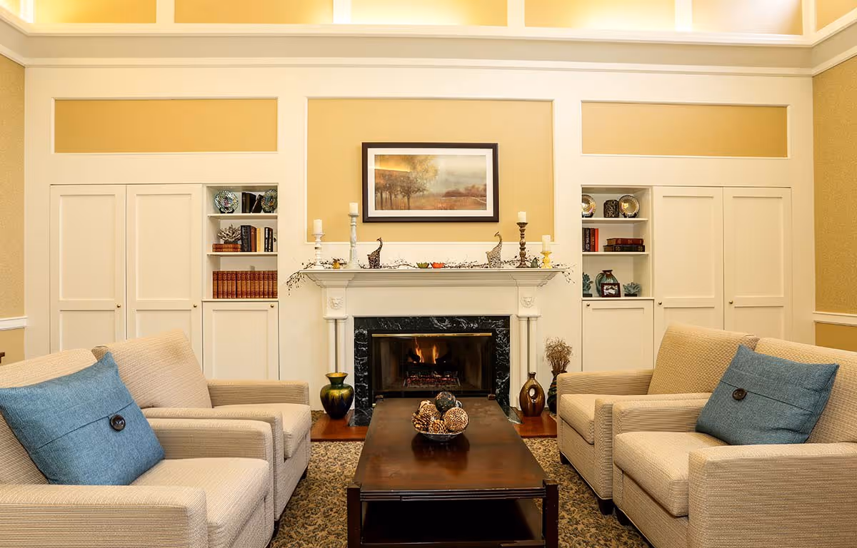A cozy living room with two beige armchairs featuring blue cushions facing a dark wooden coffee table. Behind the table is a fireplace with a black marble surround and a white mantel decorated with candles and small sculptures. Built-in white shelves and cabinets flank the fireplace, holding books and decorative items. The walls are painted a warm yellow with white trim.