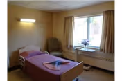 A single-bed bedroom in a senior living facility with a chair, window, and soft lighting.