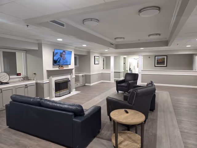 A modern senior living lounge with black leather sofas, a fireplace, and a wall-mounted TV.
