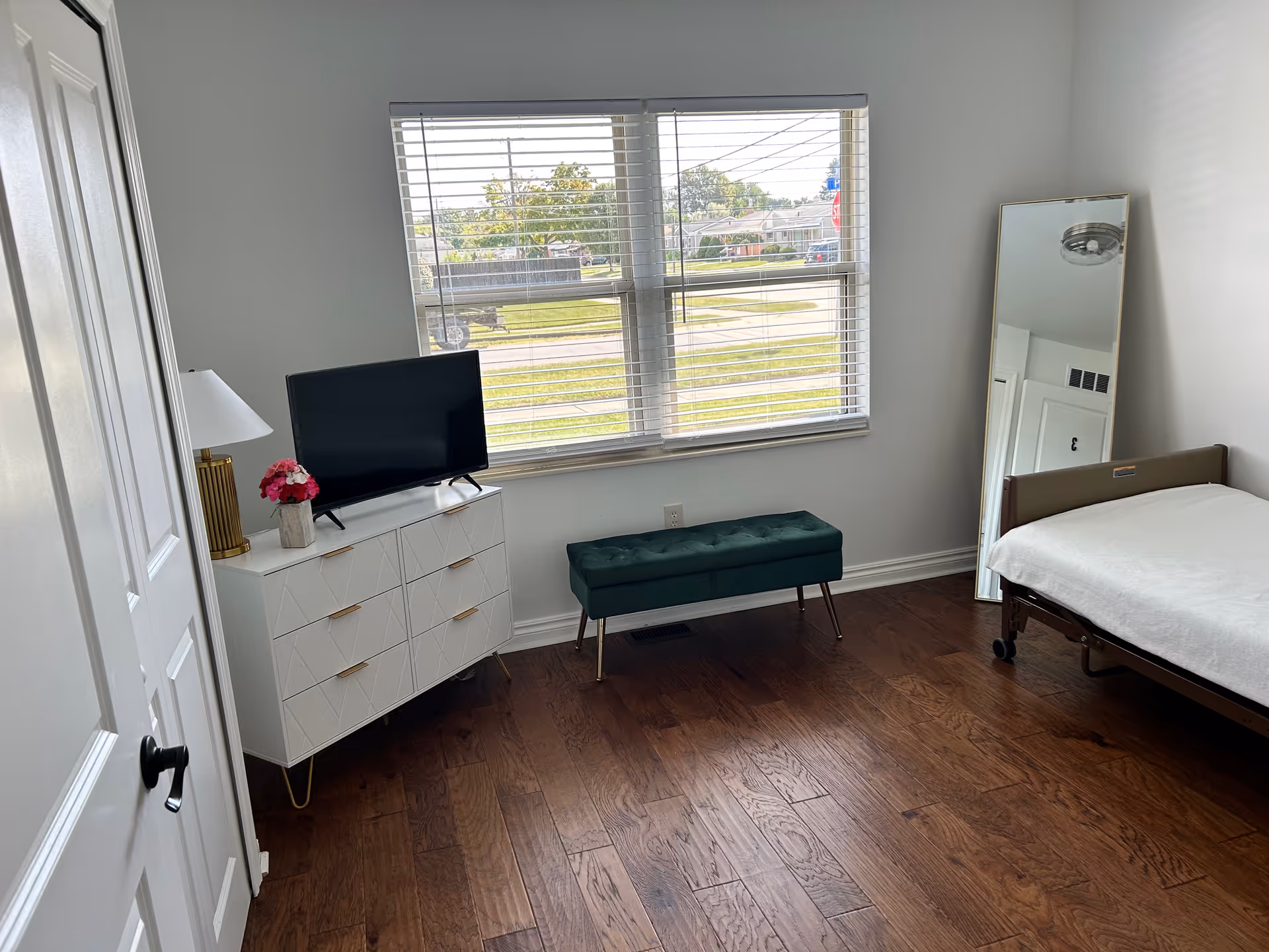 A simple bedroom with a single bed covered with a white blanket, a tall mirror leaning against the wall, a green cushioned bench under a window with blinds, and a white dresser with a TV, a lamp, and a small flower vase on top. The room has wooden flooring and white walls.