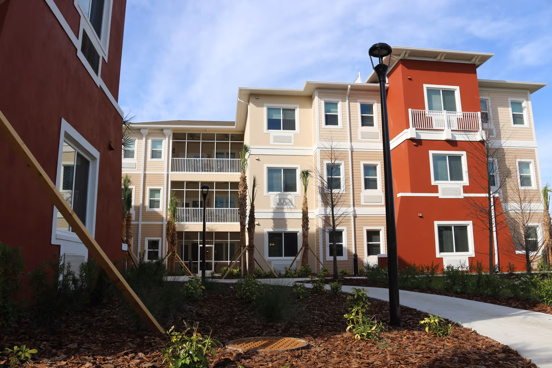 Exterior view of a multi-story senior living facility building with beige and red walls, multiple windows, balconies, young palm trees, landscaped garden beds, and a paved walkway under a blue sky.