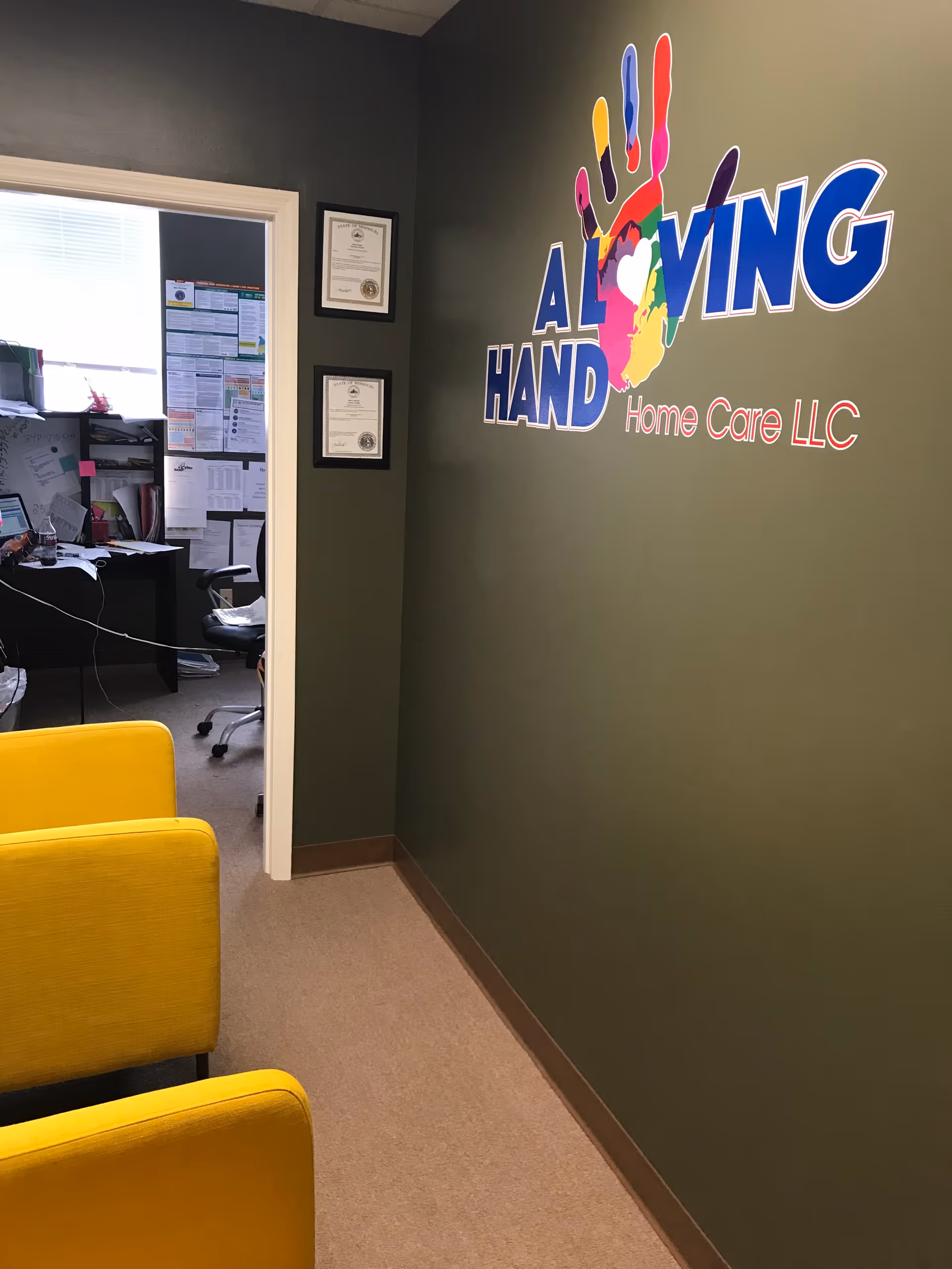 Interior hallway with office doorway, wall logo A Loving Hand Home Care LLC and yellow chairs.