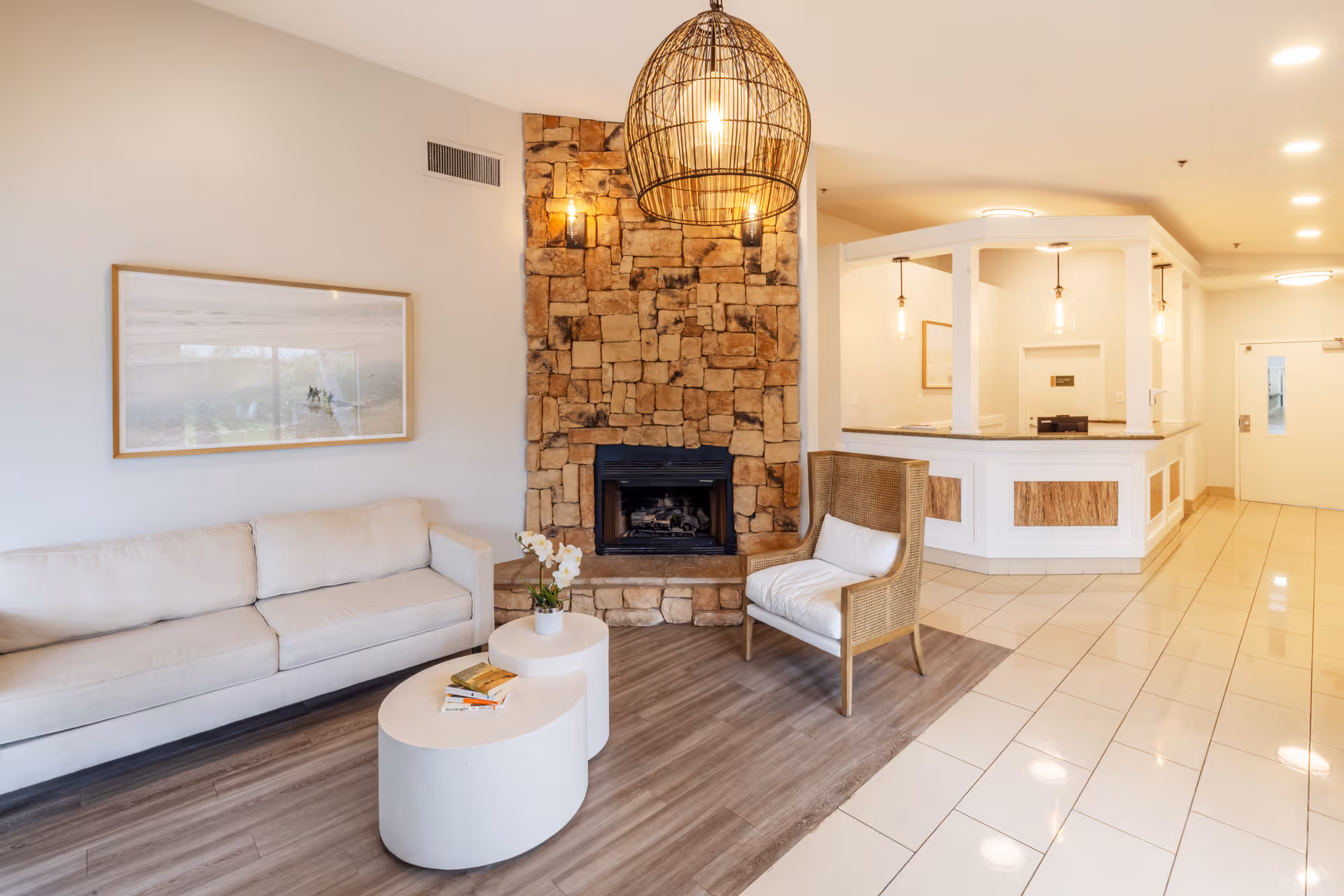 A cozy living room area with a beige sofa, a wicker armchair with a white cushion, and two white oval coffee tables with books and a small plant. The room features a stone fireplace with two wall sconces and a large woven pendant light hanging from the ceiling. In the background, there is a white reception desk with pendant lights and a tiled floor.