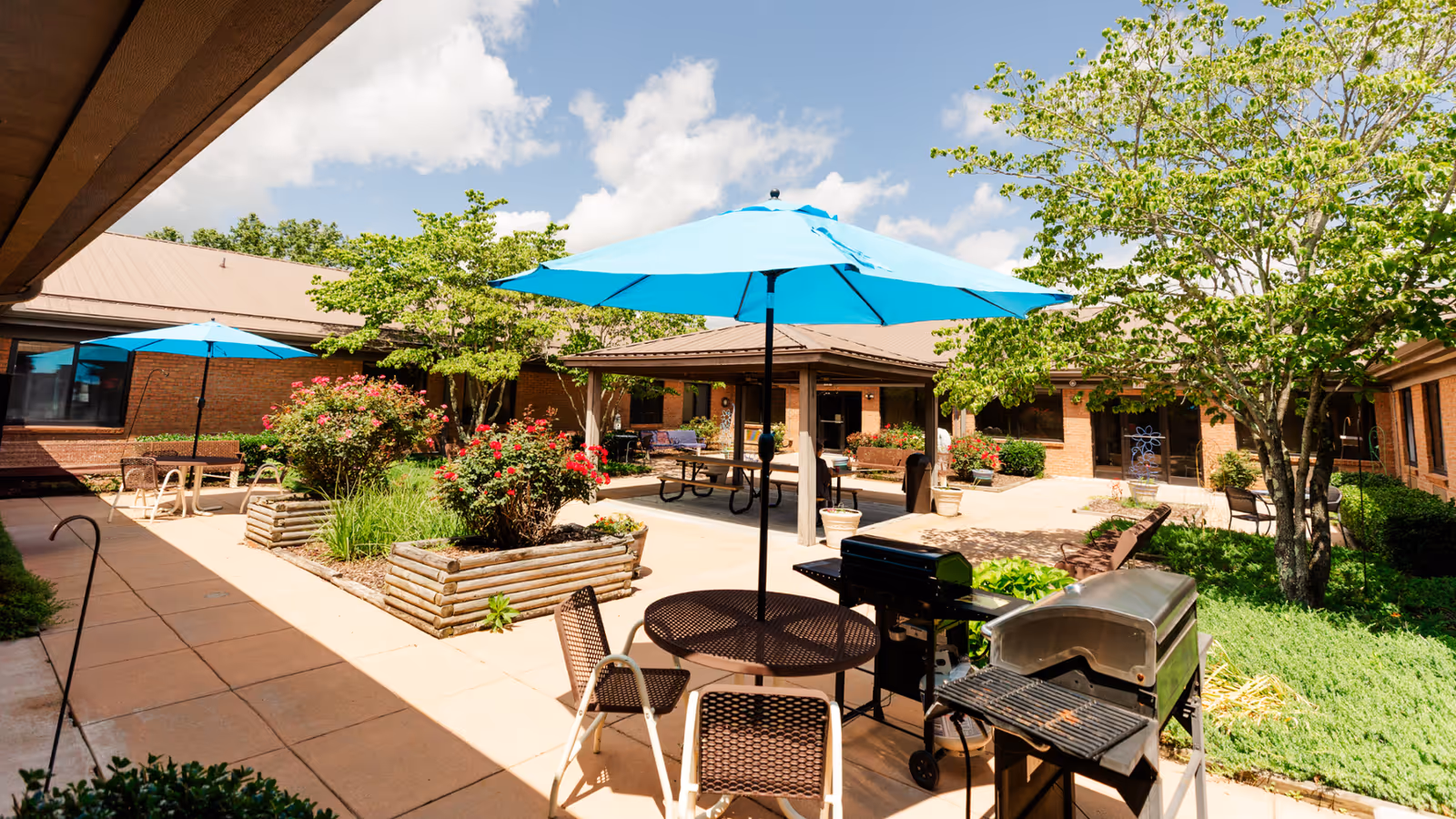 Outdoor courtyard area at Laughlin Healthcare Center featuring patio tables with blue umbrellas, chairs, a barbecue grill, raised garden beds with flowers and greenery, and surrounding brick building under a partly cloudy sky.