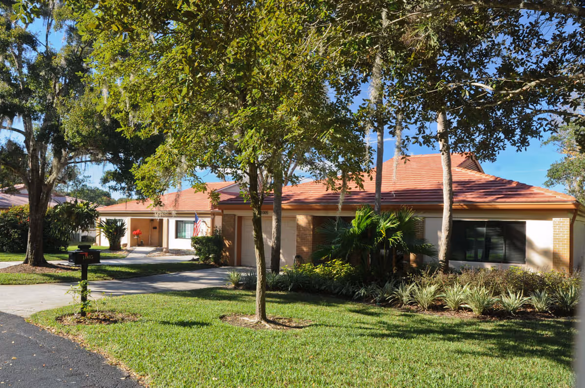 Single-story brick building with a red roof, driveway, mailbox and a grassy, tree-shaded front lawn.