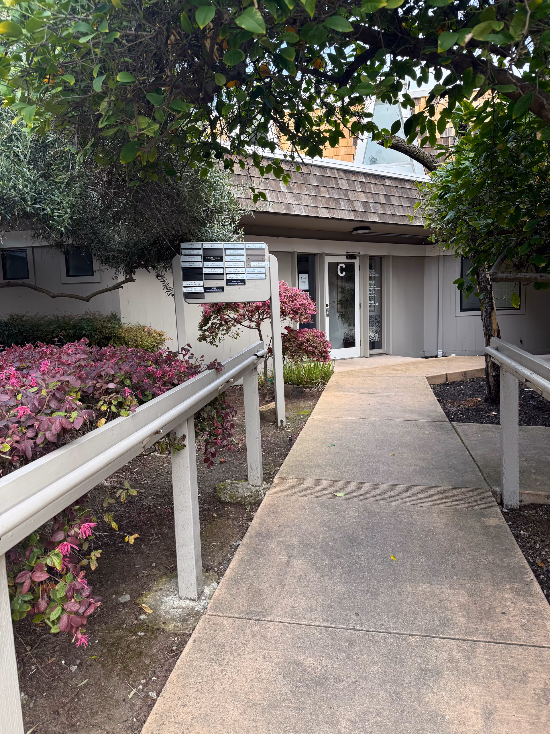 Exterior walkway to a senior living facility entrance with shrubs and a signboard.